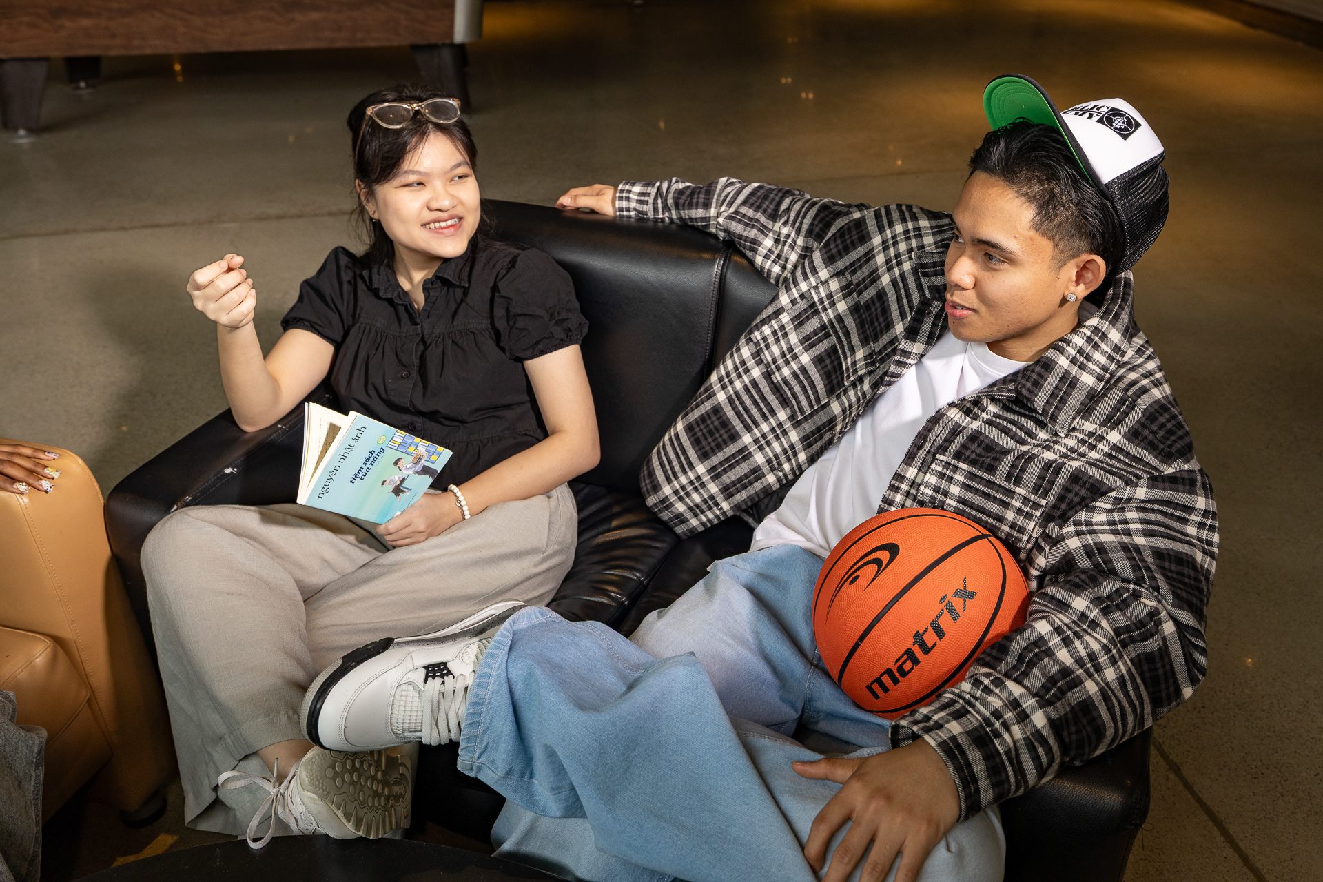 Two young people sit on black chairs indoors. The woman on the left, holding a book, talks and smiles. The man on the right, wearing a cap and plaid shirt, relaxes with a basketball on his lap and looks at her.