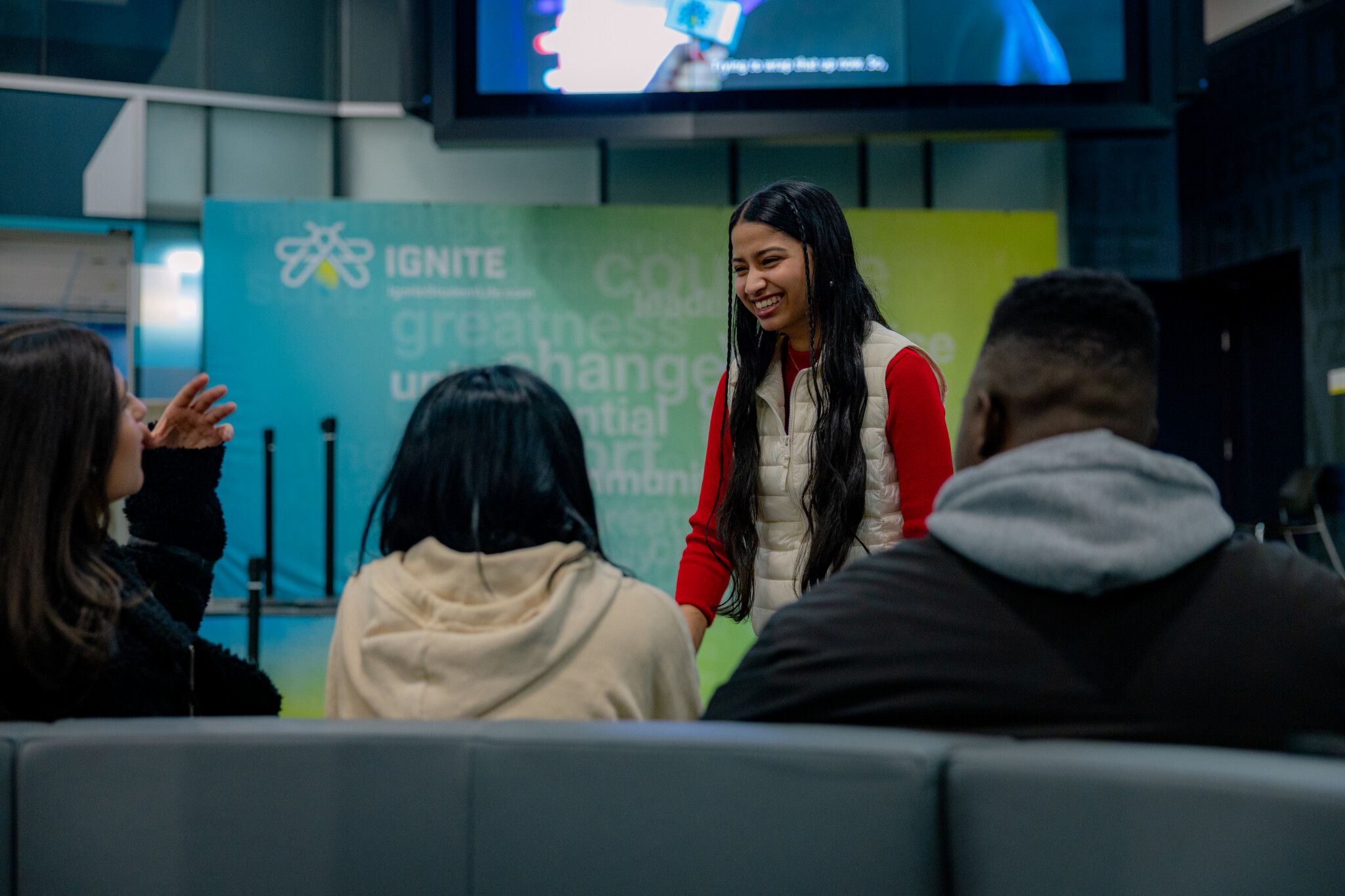 A young female student smiling and speaking with two fellow students in an engaging discussion, against the background of an IGNITE banner.