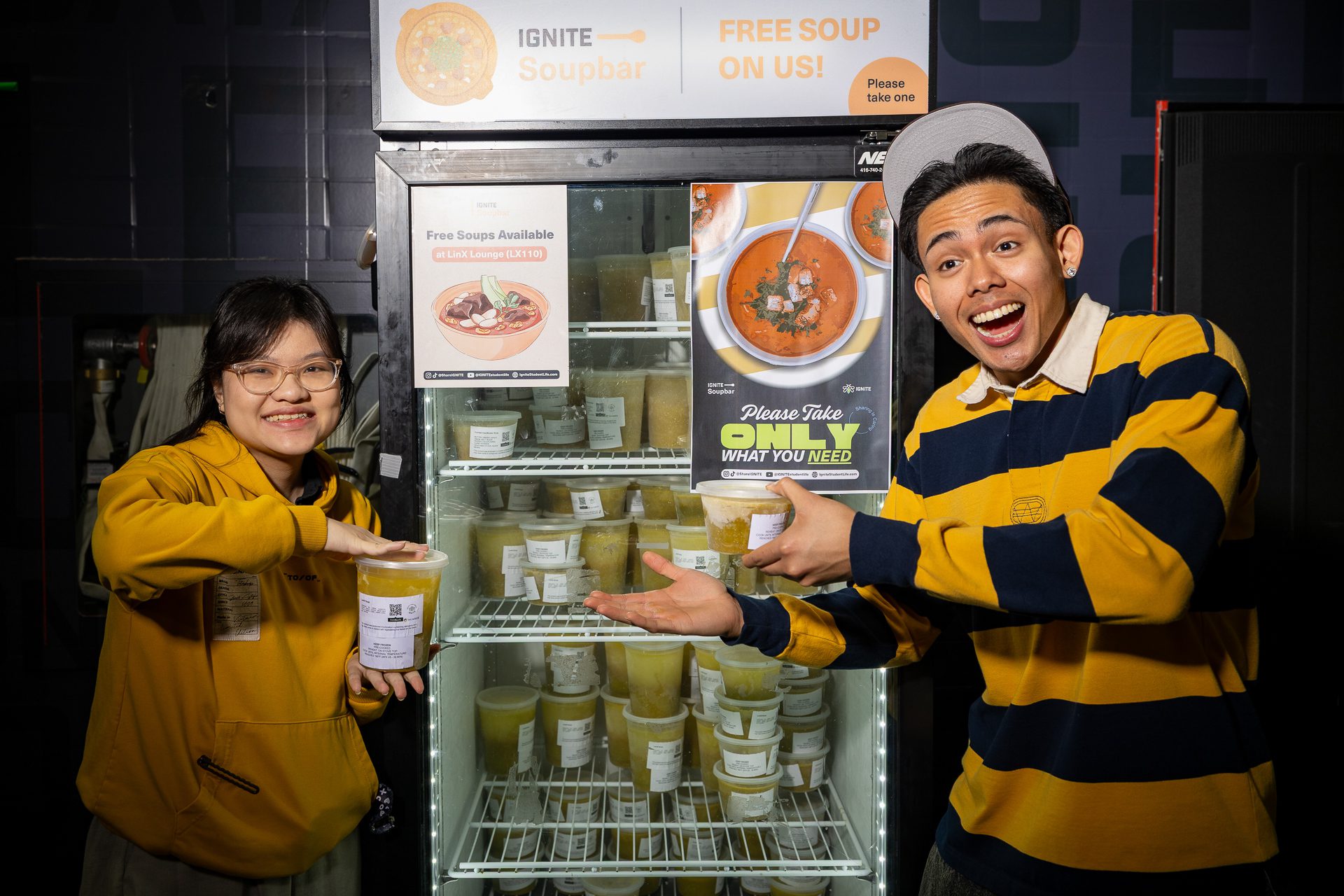 Two smiling people stand beside a fridge stocked with containers of soup. They gesture toward signs that read Free Soup and Please Take Only What You Need. Both wear yellow tops and appear cheerful.
