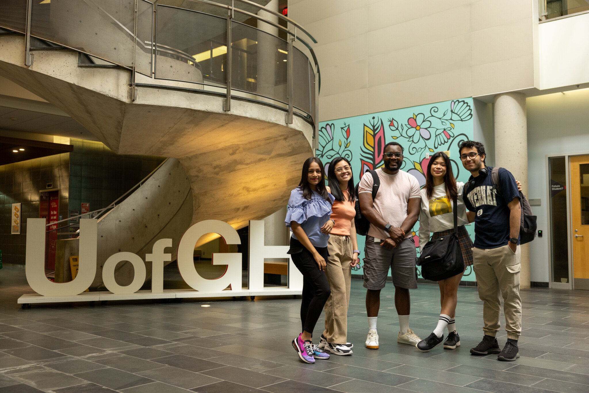 Five students stand together smiling in front of a large U of G sign inside a modern university building with a spiral staircase and a colorful mural in the background.
