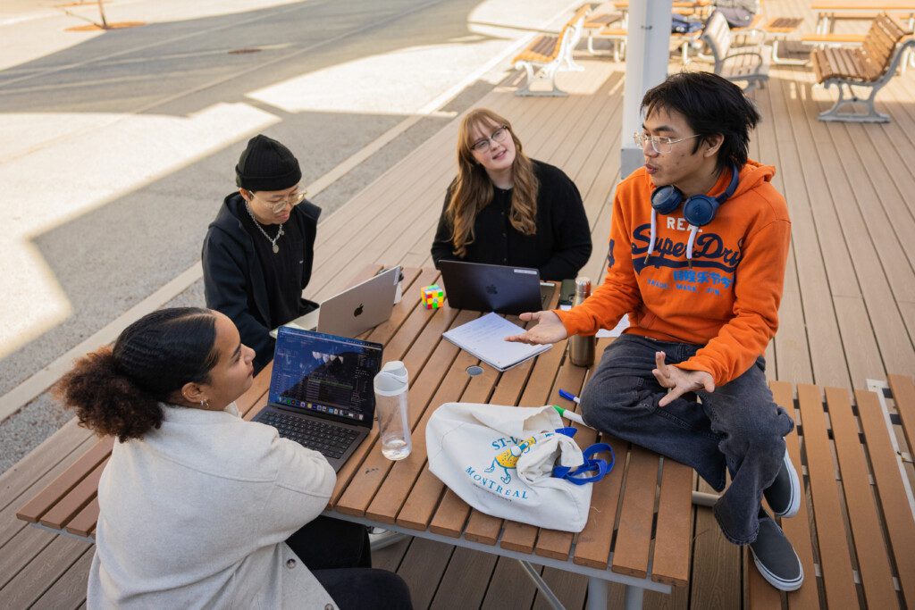 Four students seated at an outdoor picnic table with laptops, listening to a peer in an orange hoodie speak.