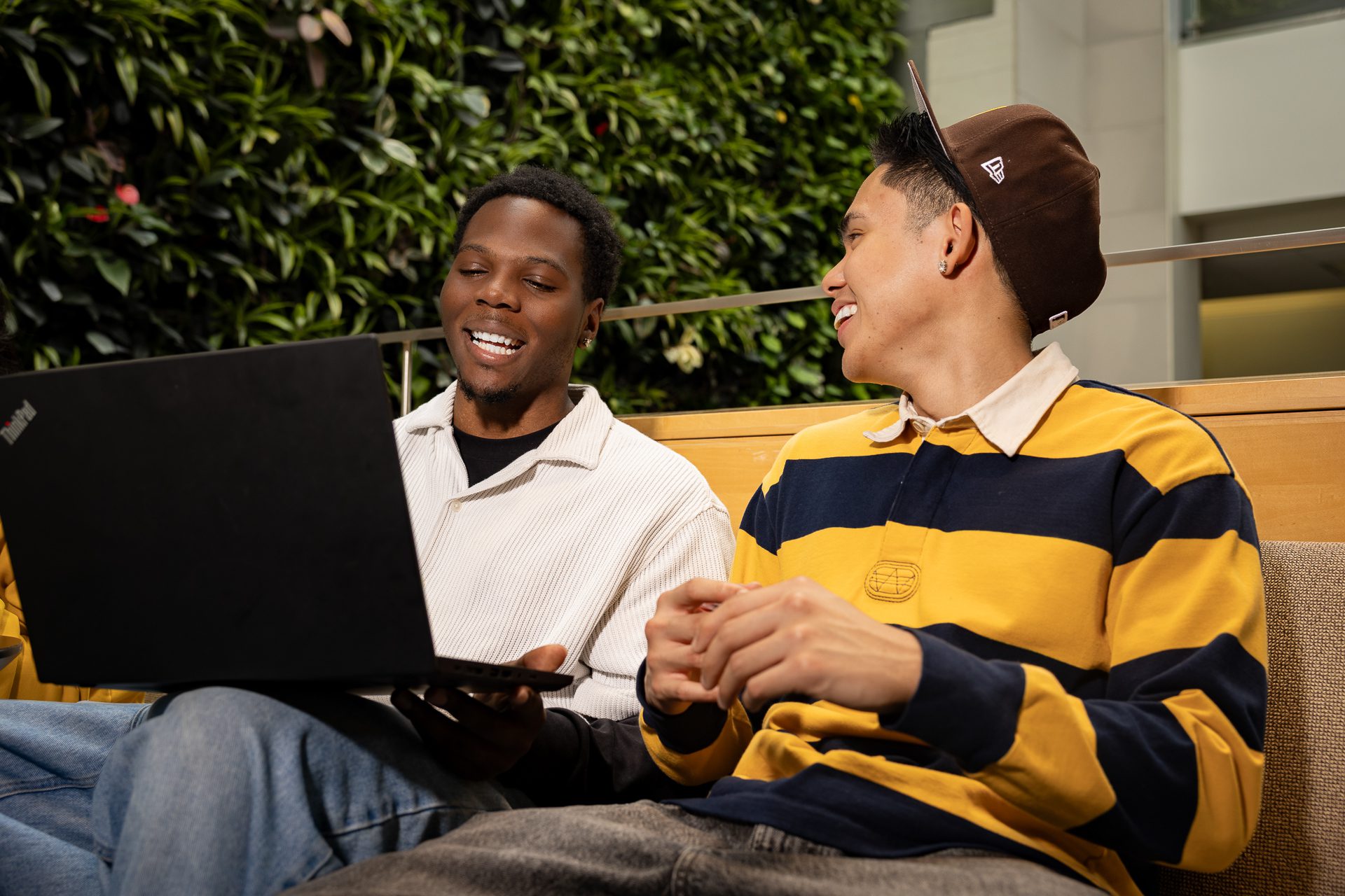Two young men sit on a bench indoors, smiling and talking while looking at a laptop. One wears a beige shirt and jeans, the other wears a brown cap and a yellow-striped shirt. A green wall is in the background.