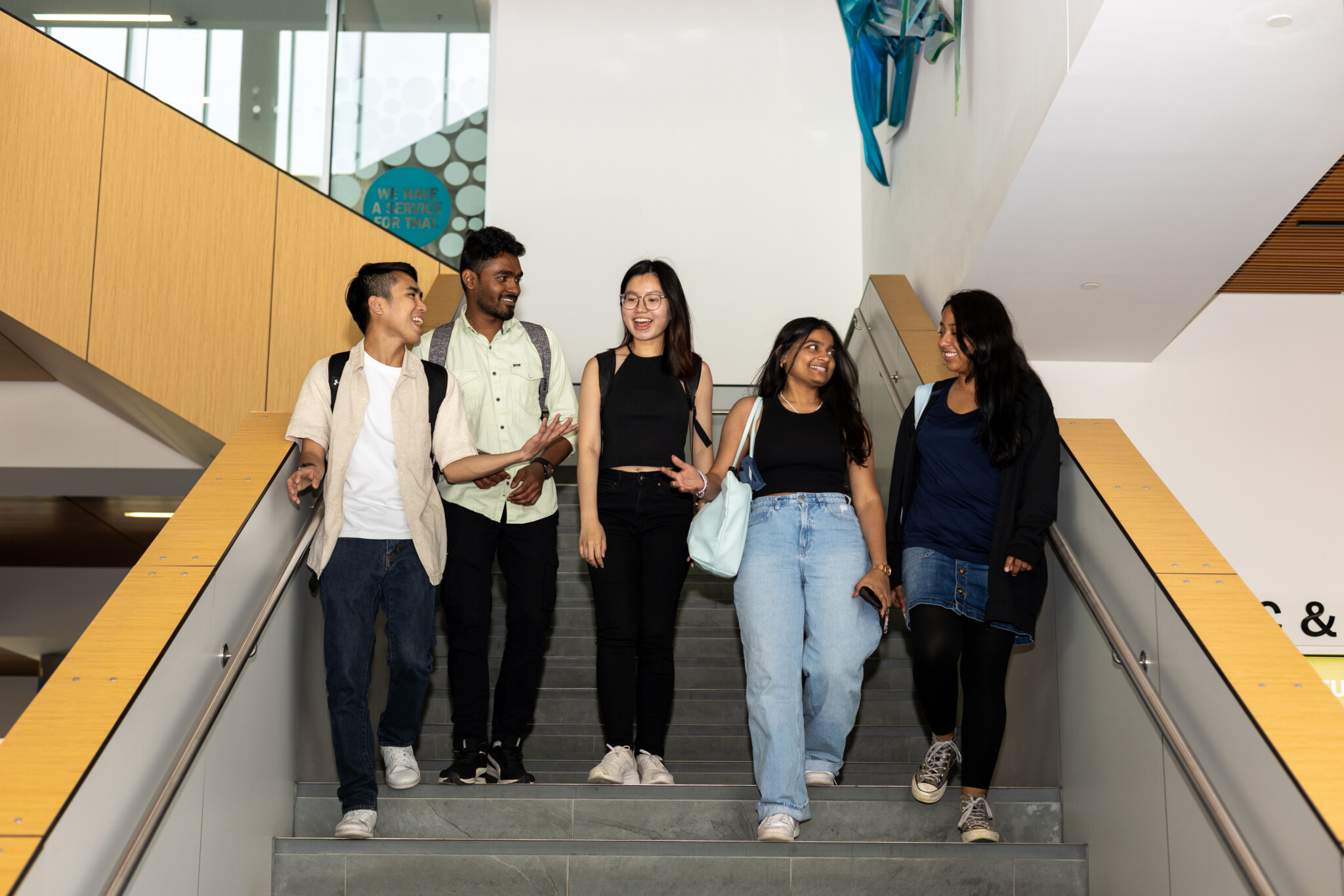 Five young adults walk down a staircase indoors, smiling and talking. They appear to be students, carrying backpacks and bags, and are casually dressed. The background shows a modern building interior.
