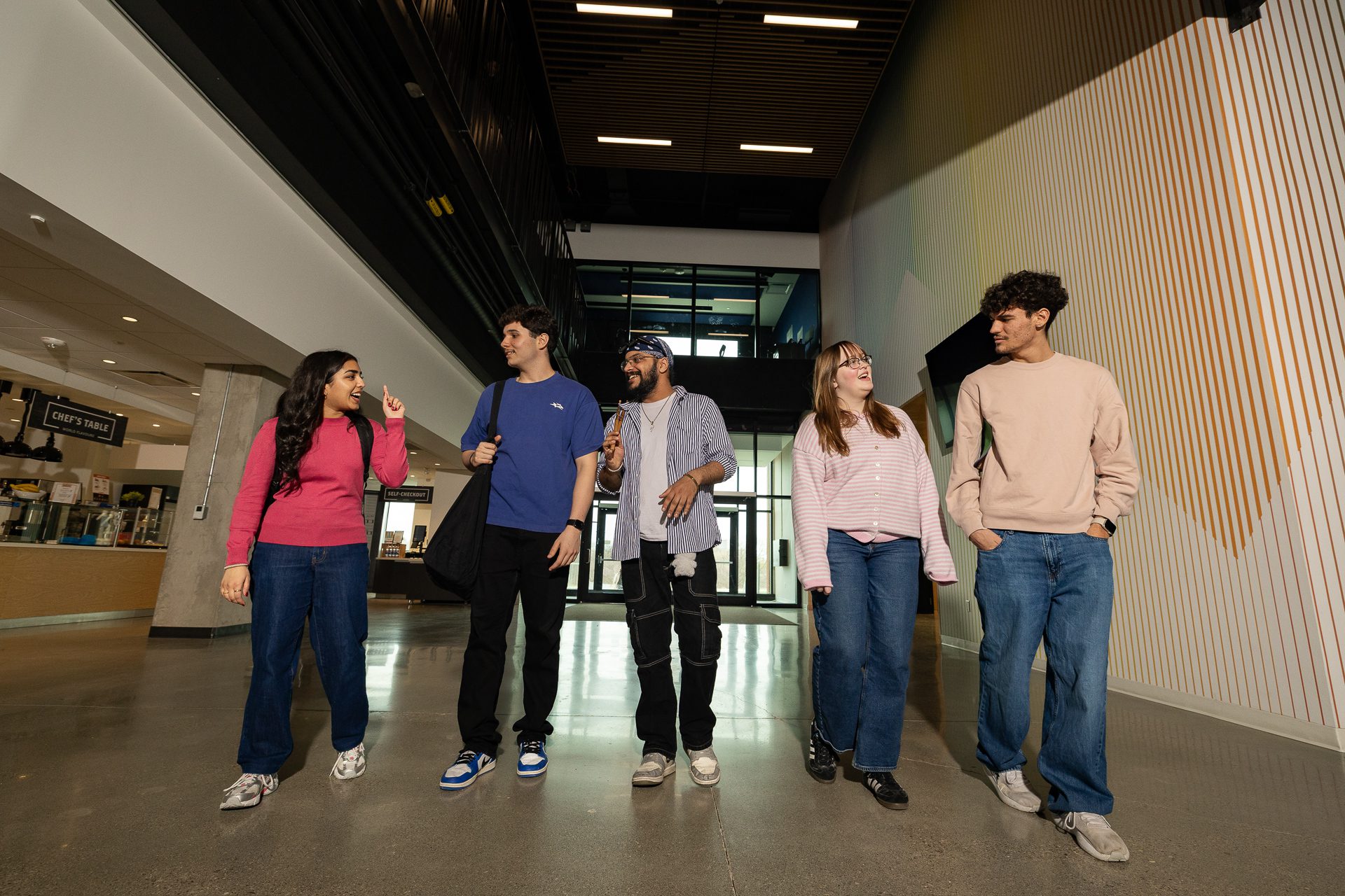 Five students walk together in a modern school hallway, talking and smiling. They wear casual clothes and appear to be engaged in friendly conversation. Natural light comes in through windows behind them.