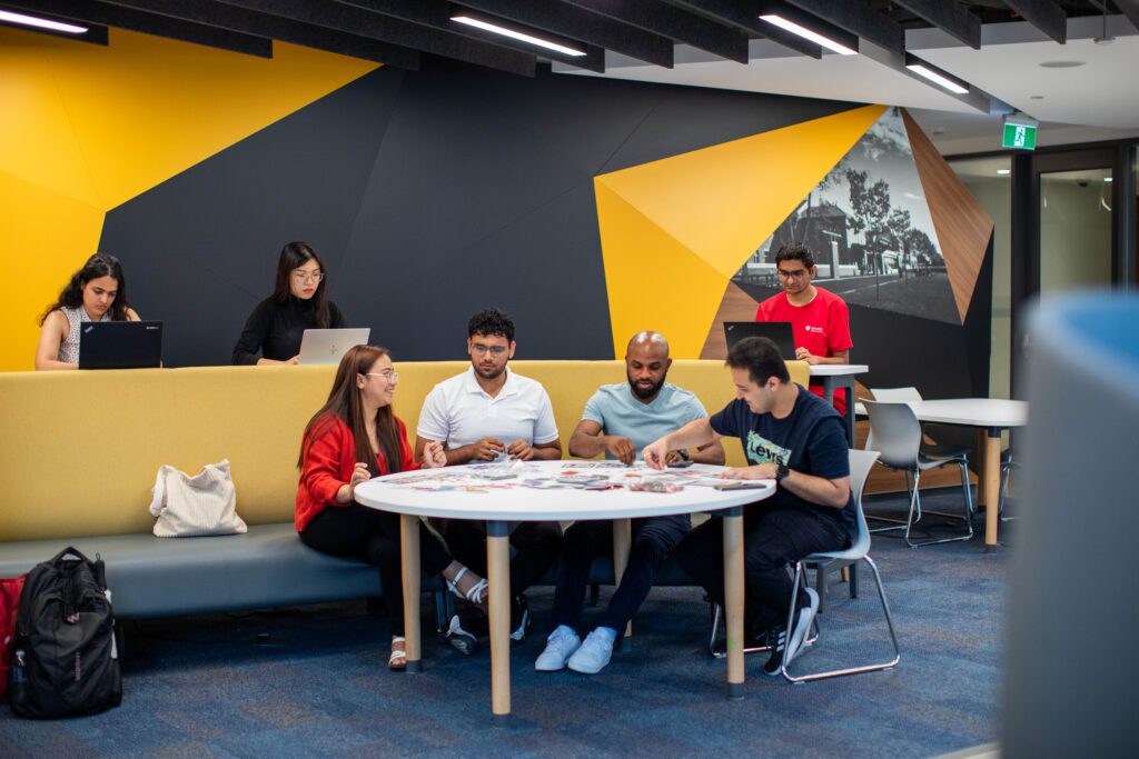 A group of five people sit around a round table, looking at cards or papers, while two others work on laptops at a nearby bench in a modern, brightly colored lounge or study area.