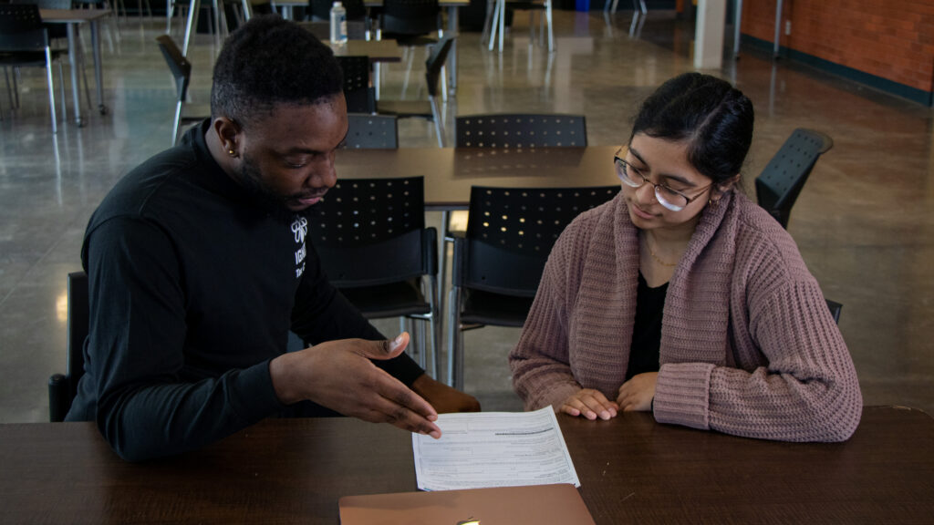 Two people sit at a table in a spacious room, looking over a document together. One person gestures toward the paper while the other listens and follows along attentively.