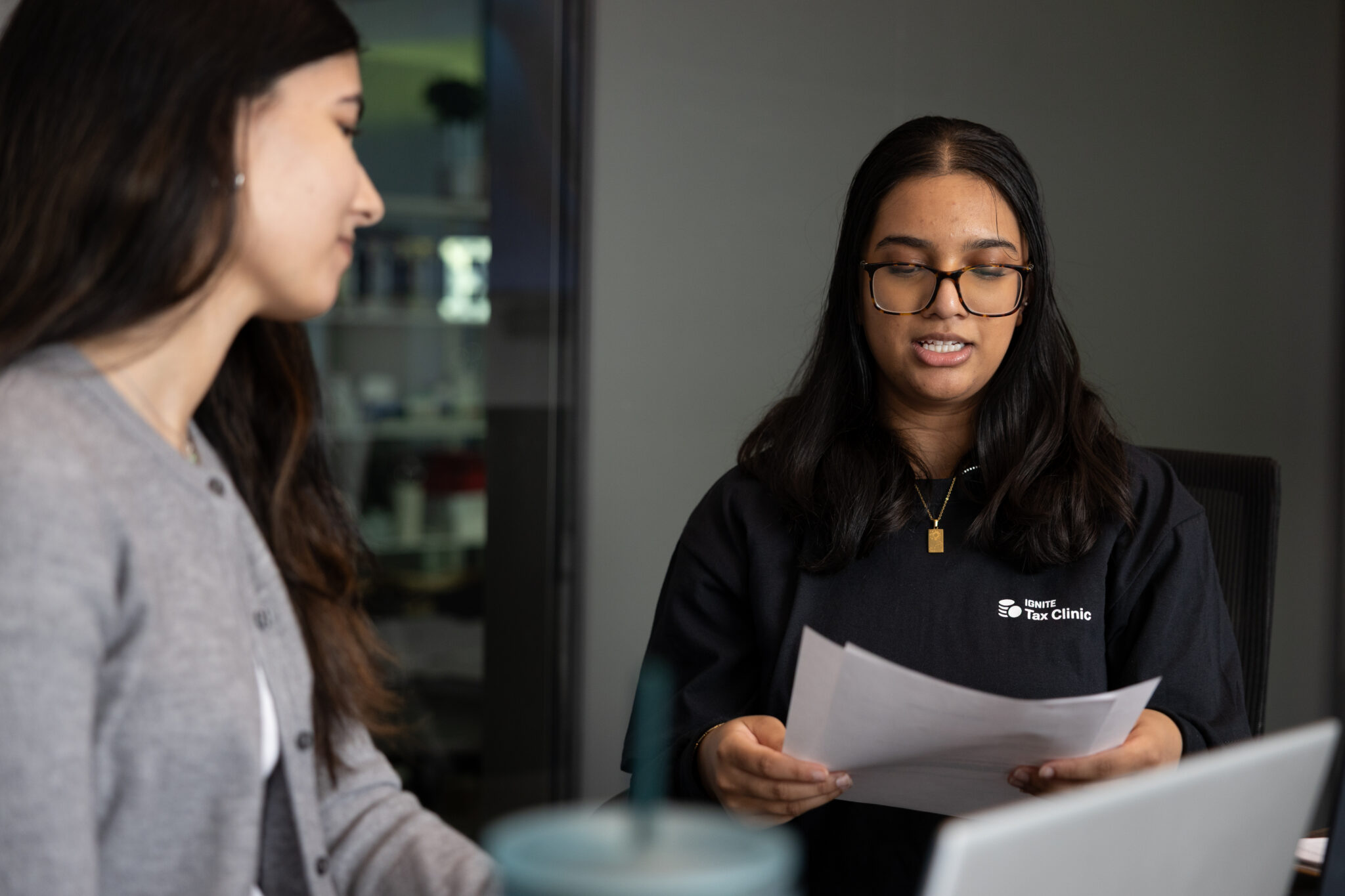 Two women sit at a table indoors. One woman, wearing glasses and a black “Tax Clinic” shirt, holds and looks at paperwork while speaking. The other woman listens, partially visible, facing her.