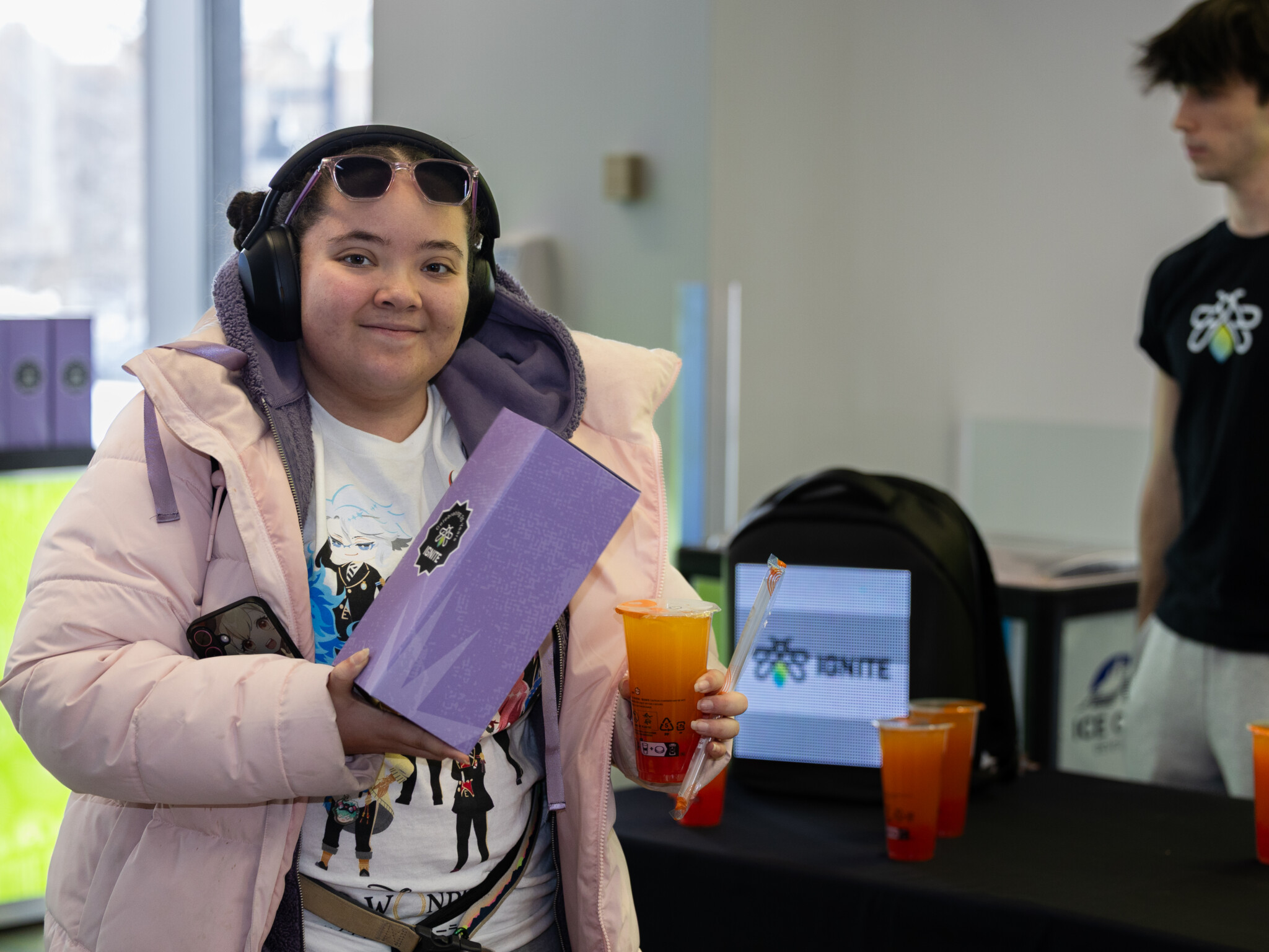 A person in a light purple coat holds a purple box and a drink, wearing headphones and smiling. Another person stands nearby, and bubble tea drinks are on a table with an IGNITE sign in the background.