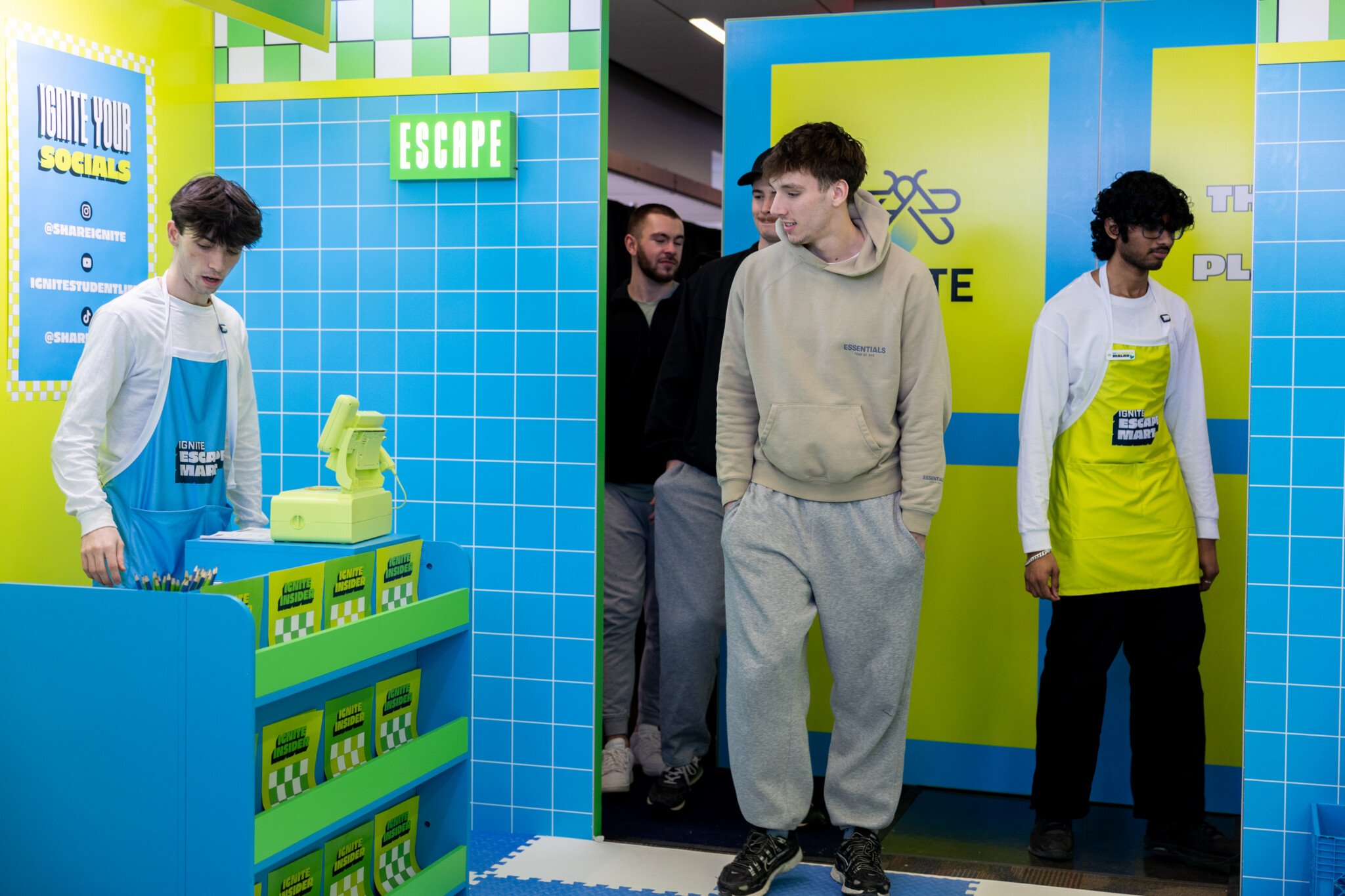 Group of young men at a colorful booth with blue tiles and bright yellow panels; a staff member in a blue apron stands at a neon counter with pamphlets nearby.