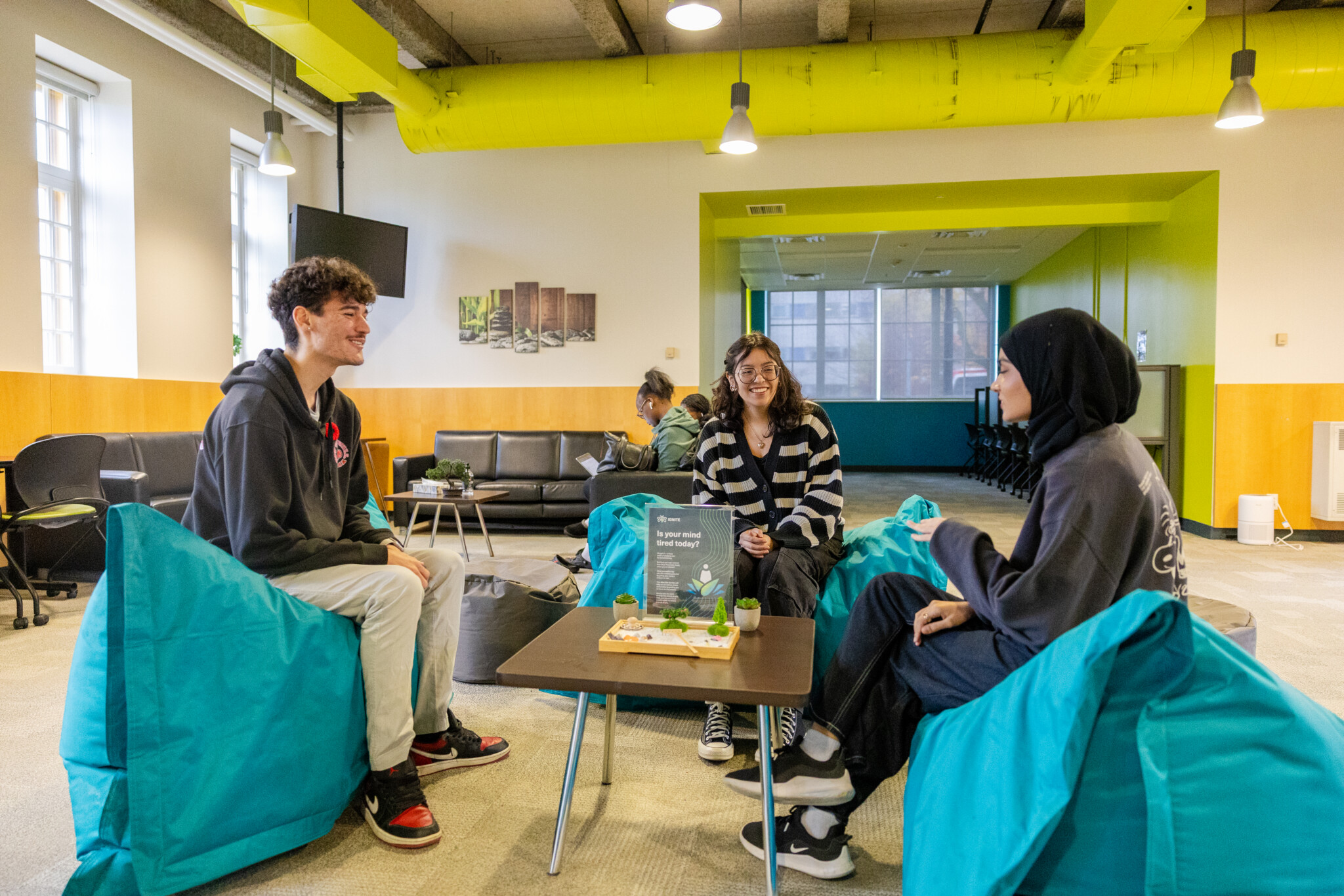 Three students chatting while seated on large turquoise beanbag chairs in a modern student lounge area.