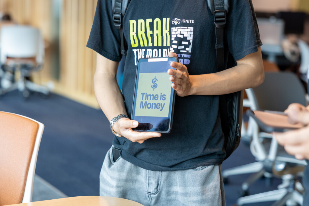 A person holds a tablet displaying the phrase Time is Money with a clock and dollar sign. The person wears a backpack and a BREAK THE MOULD t-shirt.