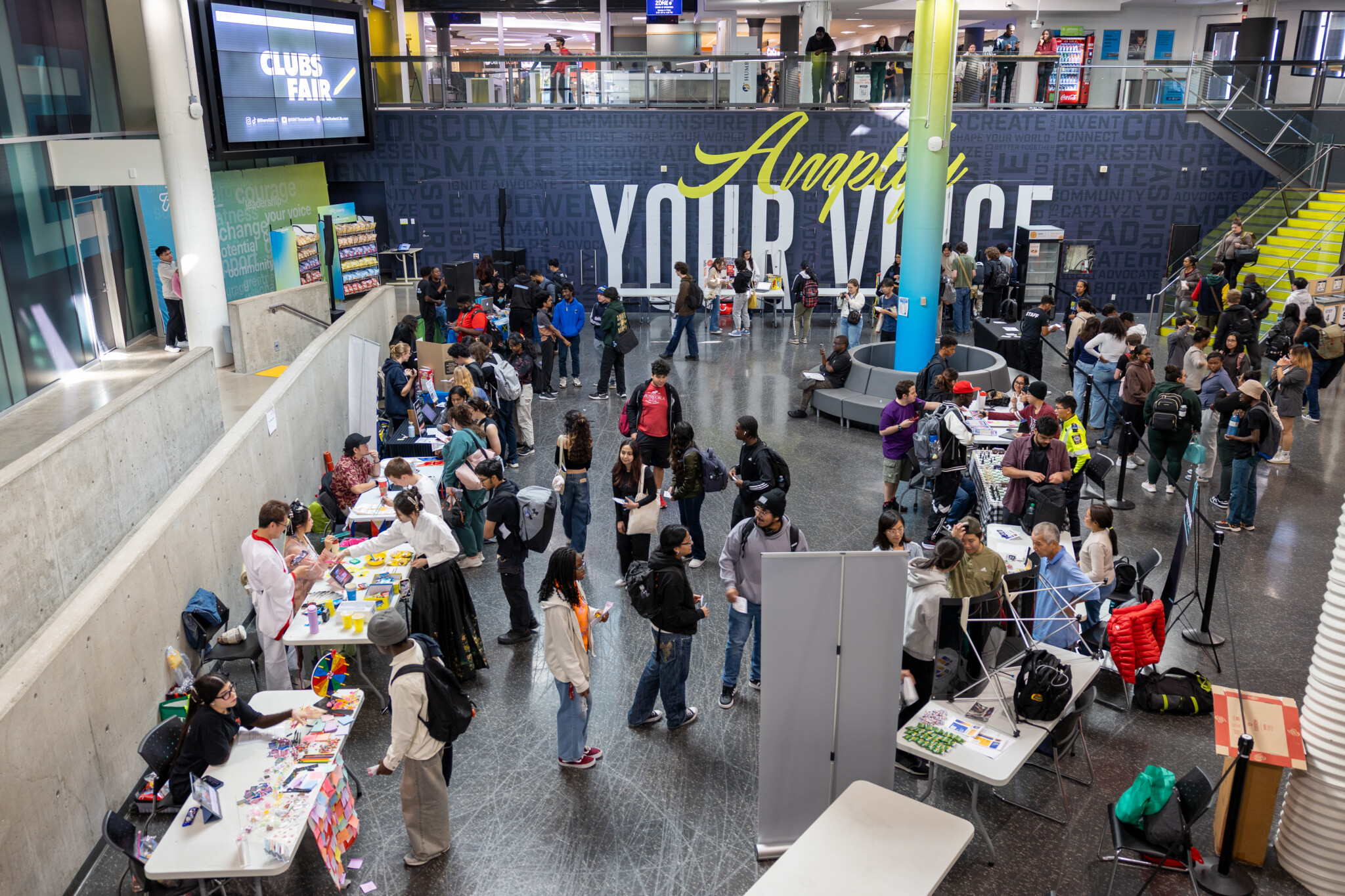 A busy indoor college clubs fair with students visiting tables, talking, and picking up flyers. Signs and posters are visible, and a large wall reads “Amplify YOUR VOICE.” Bright lighting and stairs are in the background.