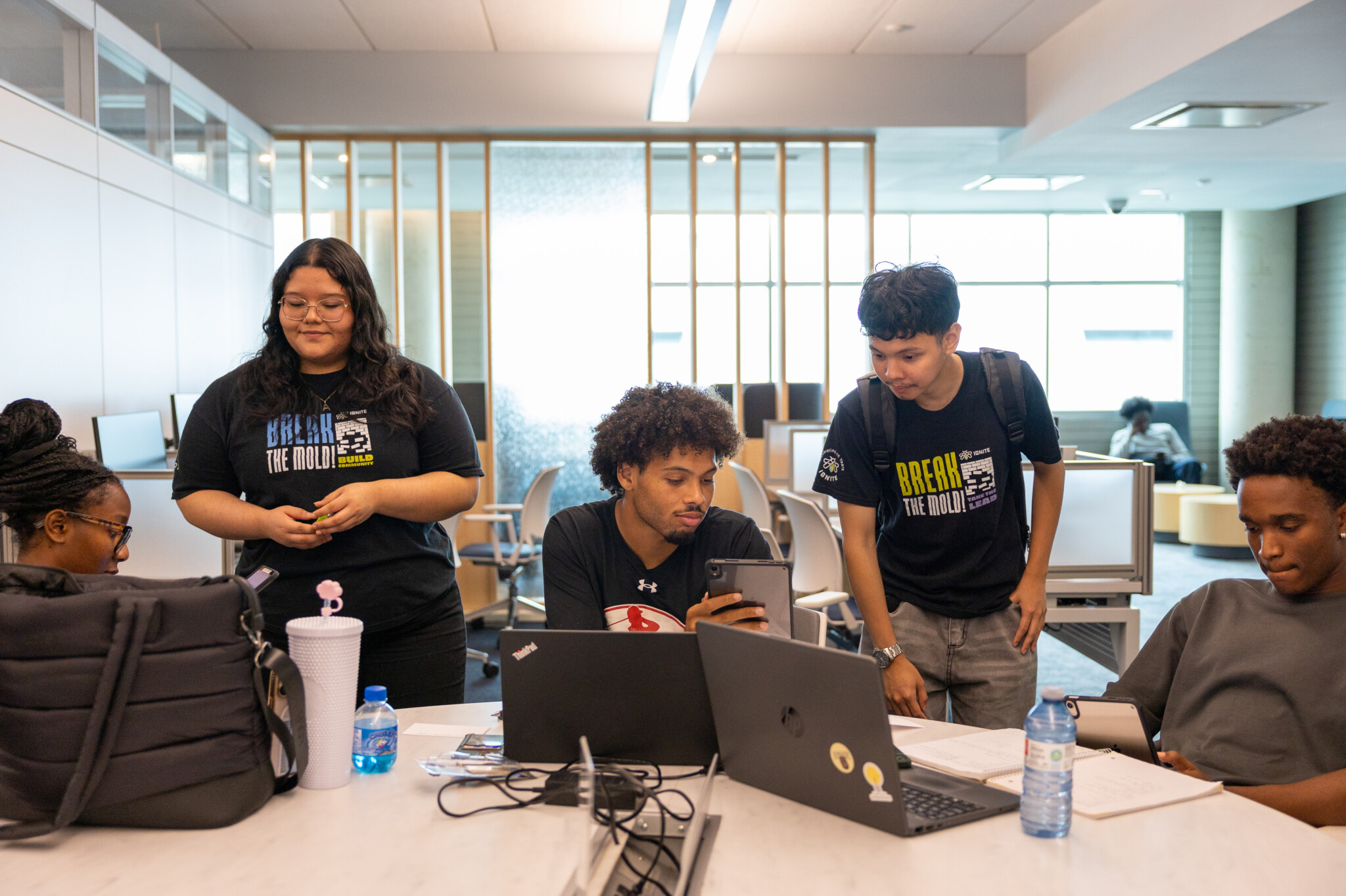 Five students work together at a table with laptops, notebooks, water bottles, and bags in a bright, modern study area. Two students stand while three sit and look at a phone.