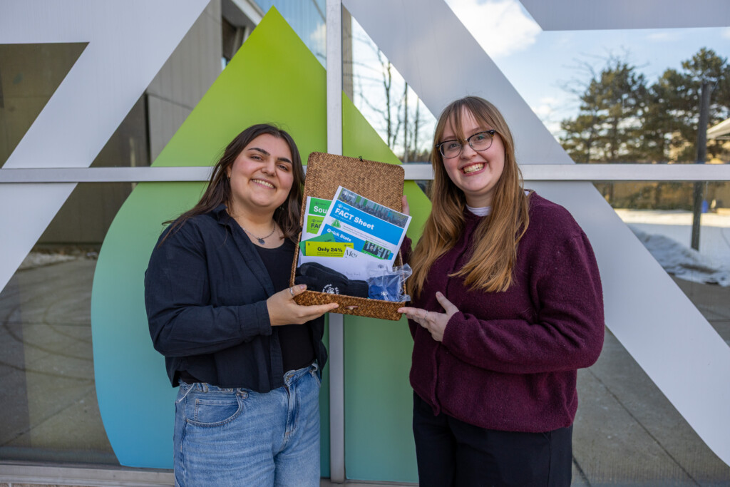 Two people smiling outside, holding an open box containing pamphlets, a toy car, and other items. Geometric window art and snowy ground are visible in the background.