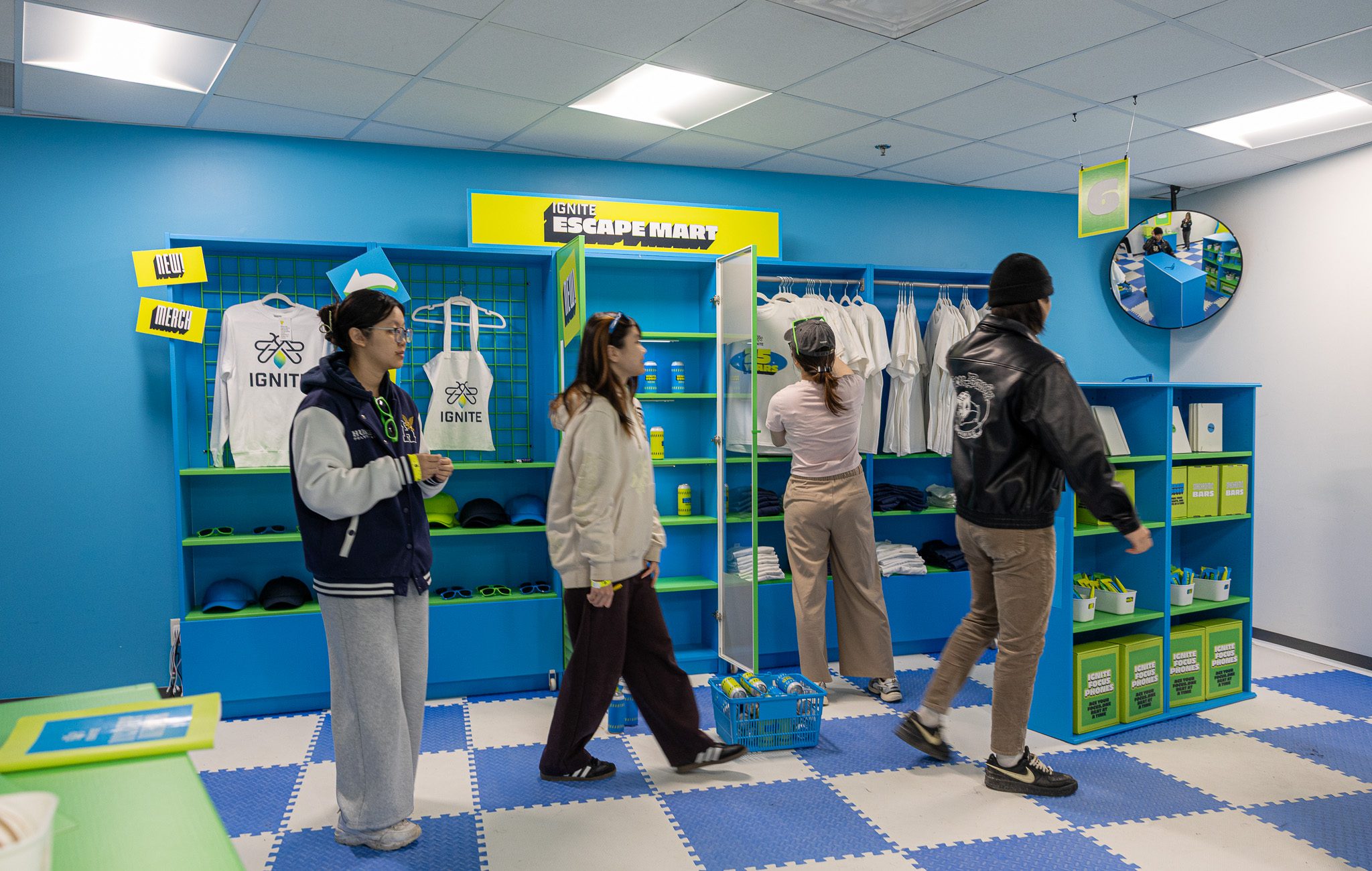 Four people stand and walk in a blue-themed store with shelves of white shirts and green bins. One person carries a shopping basket, while another appears to look at items on a shelf. A round mirror hangs in the top right corner.