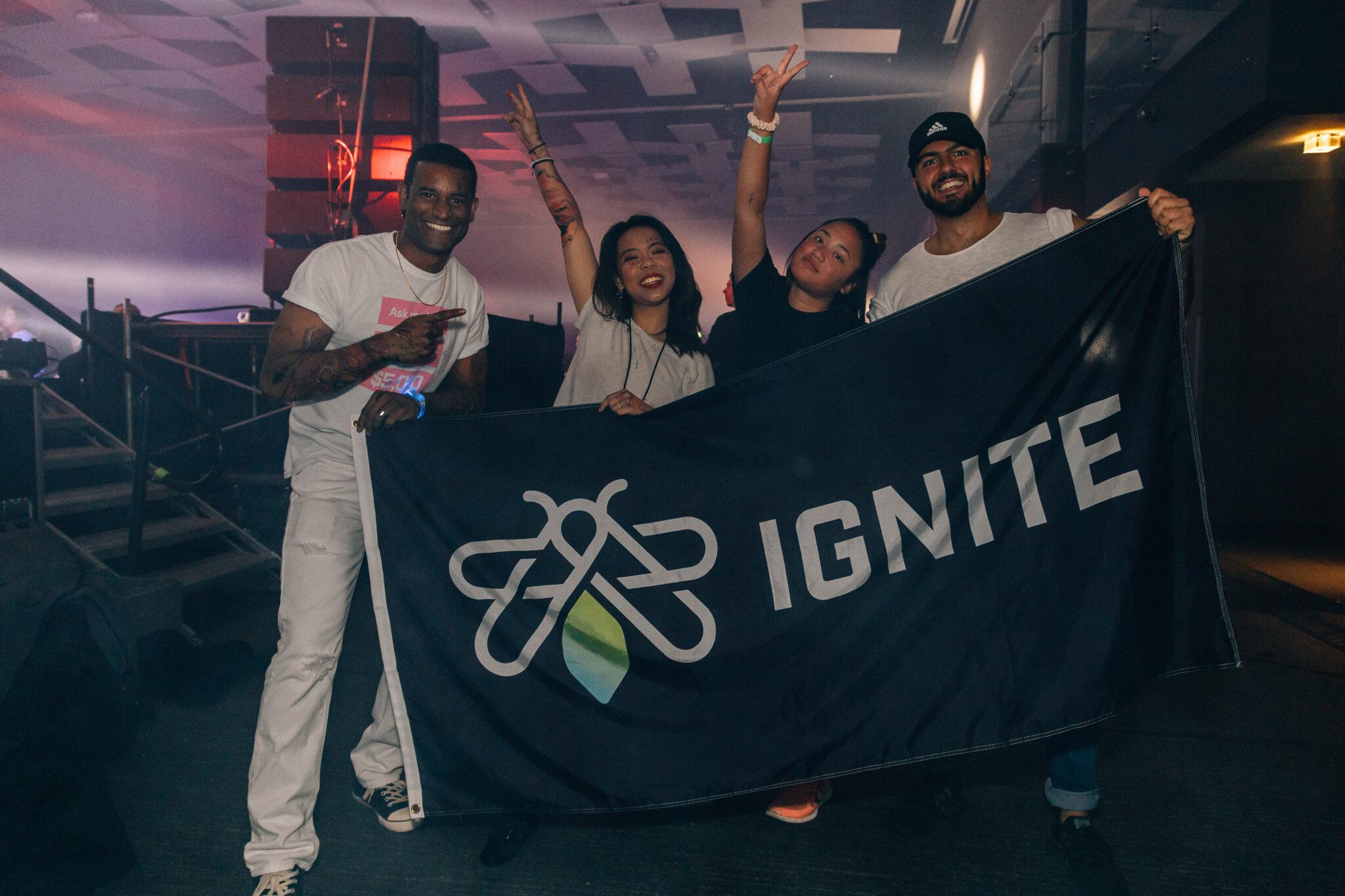 Four people smiling and making peace signs while holding up a black IGNITE flag.