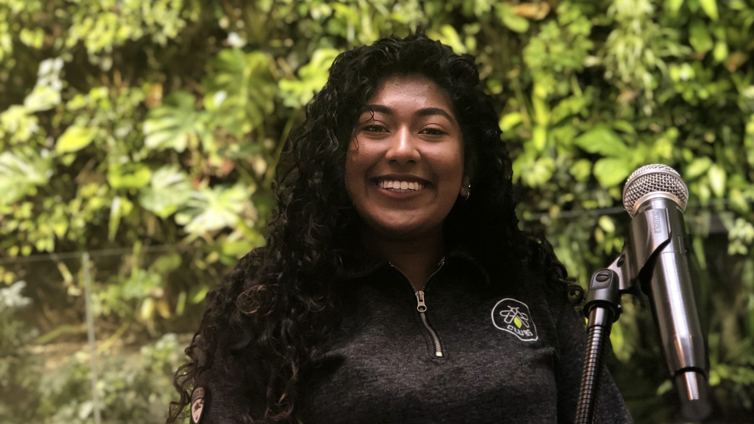 A woman with long curly hair stands smiling in front of a microphone, wearing an IGNITE sweater, with a living green wall behind them.