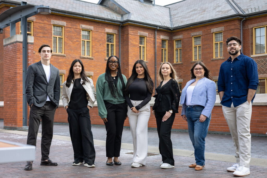 Seven student members of the IGNITE Board of Directors standing together outdoors in front of a red-brick university building.