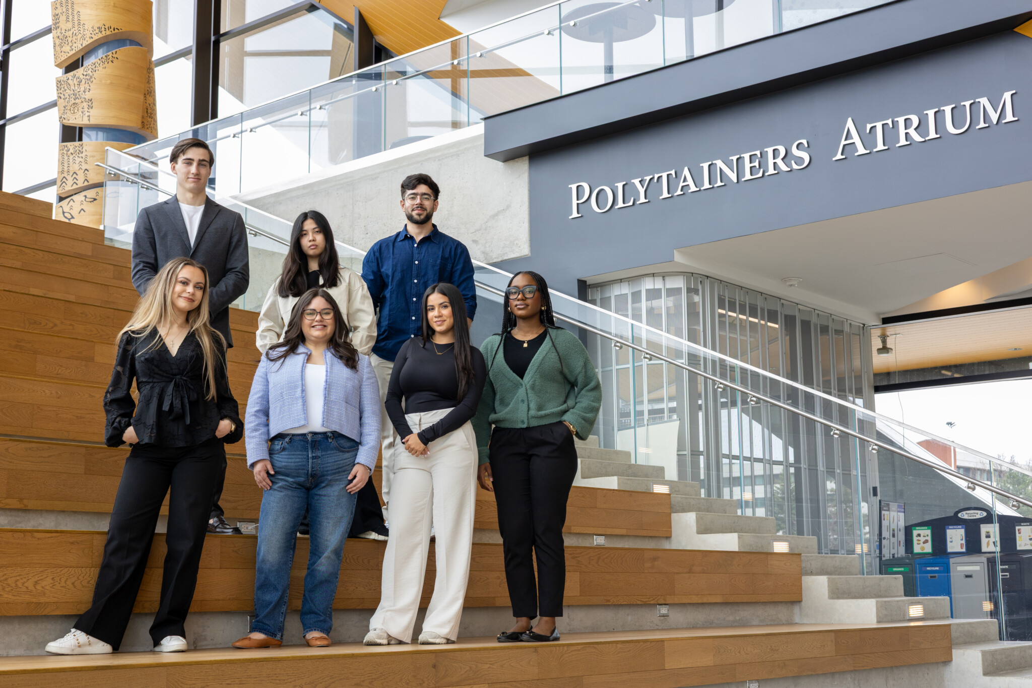 Seven people stand and sit on wooden steps inside a modern building, near a sign that reads Polytainers Atrium. They are posing for a group photo and appear to be dressed for a professional or academic environment.