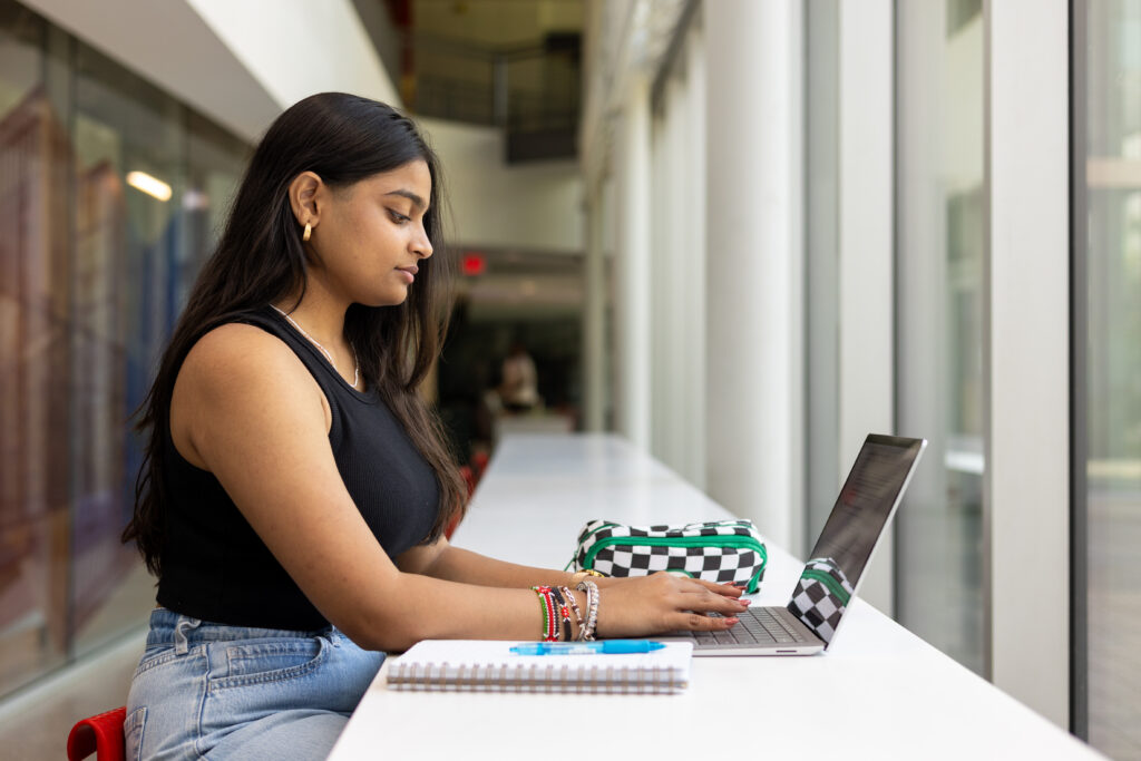 A female student typing on a laptop at a long white desk next to large windows on campus.