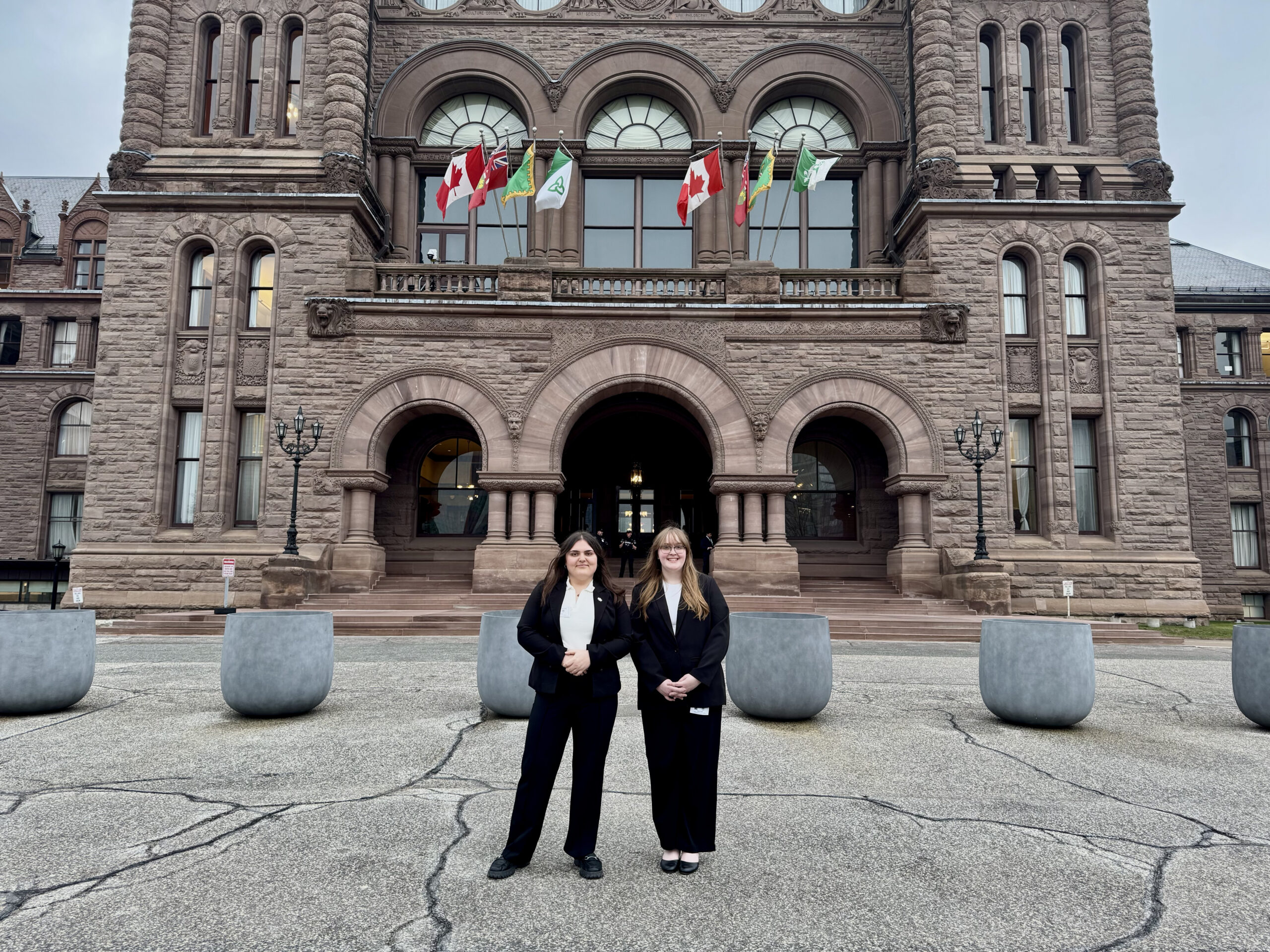 Two people in business attire stand in front of Queen’s Park.