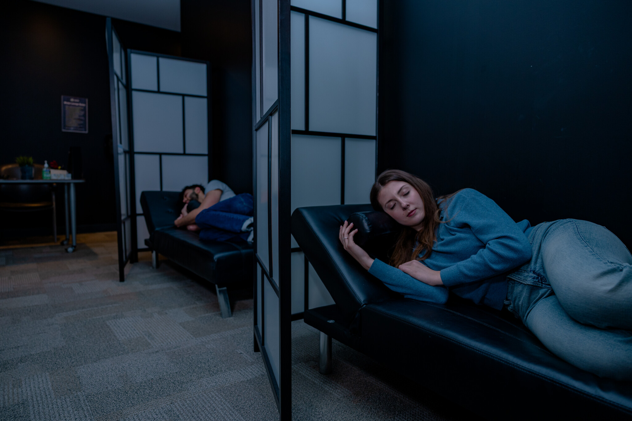 Two people lie on separate black couches in a dimly lit room with dark walls and privacy screens, appearing to rest or nap.