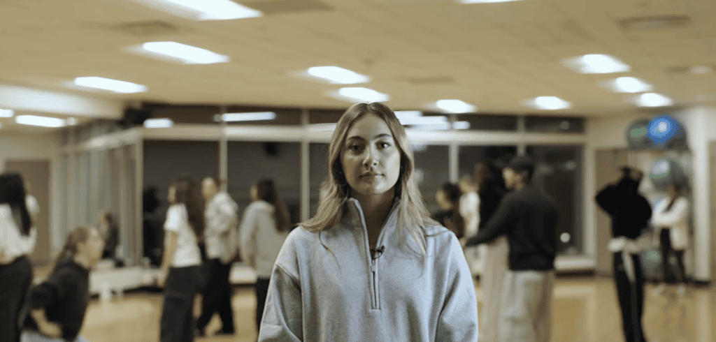 A young woman in a grey quarter-zip sweater standing in focus inside a indoor fitness studio with people in the background.