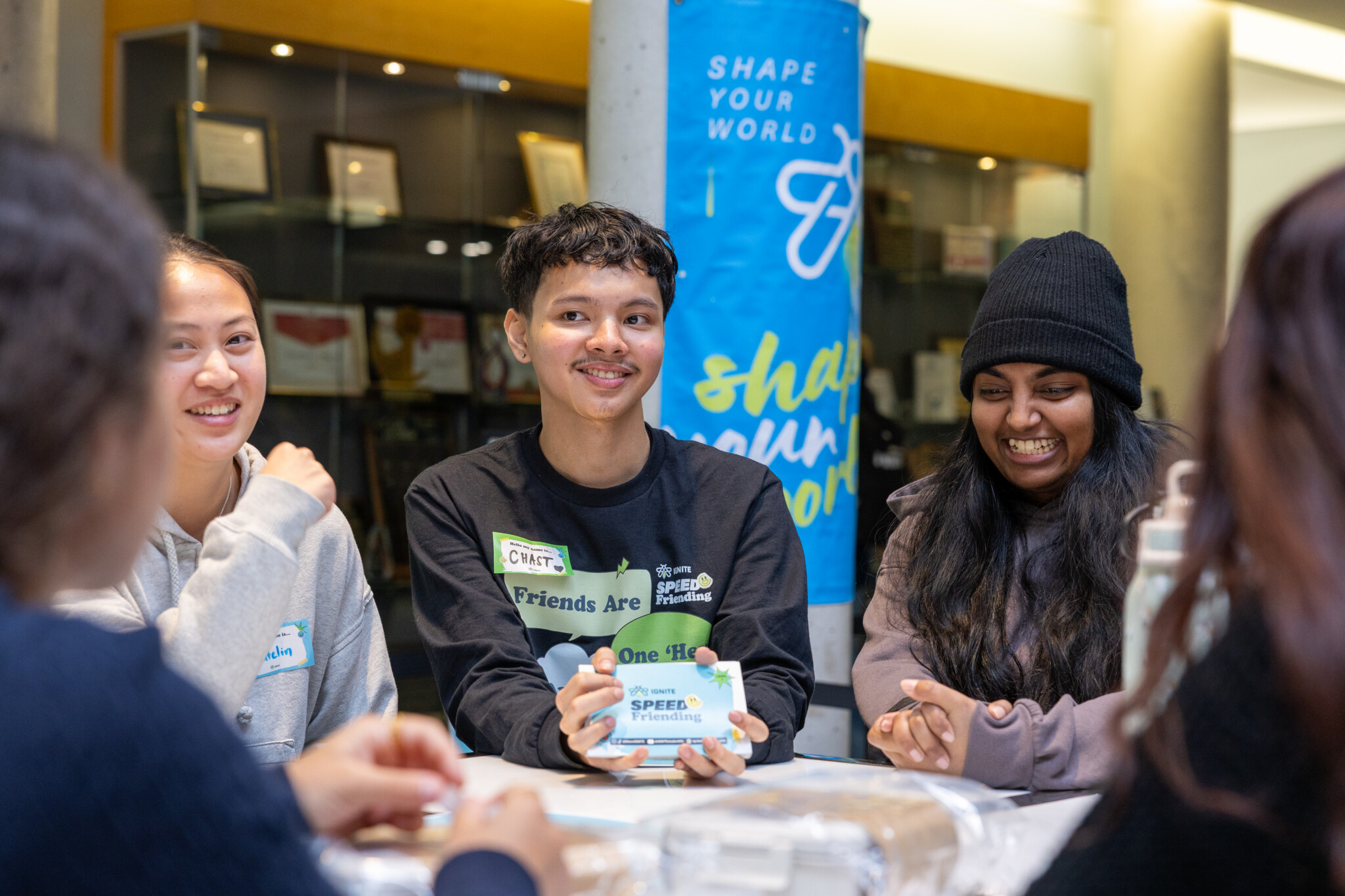 Group of smiling young people around a table indoors, sharing cards and chatting, with a bright blue banner in the background reading 'Shape Your World'