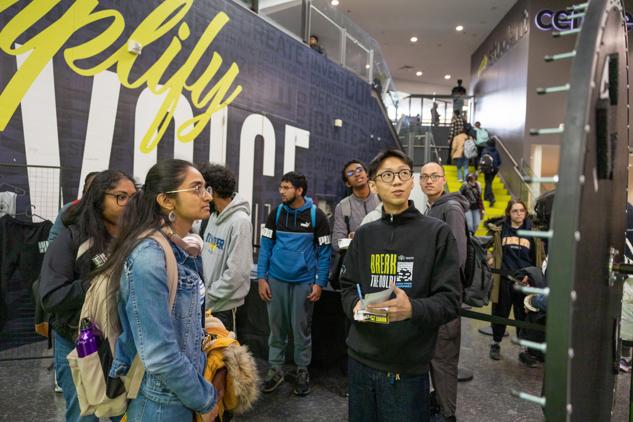 Group of students in a busy hallway at a tech/event, with a yellow mural on the wall behind them. (informative)
