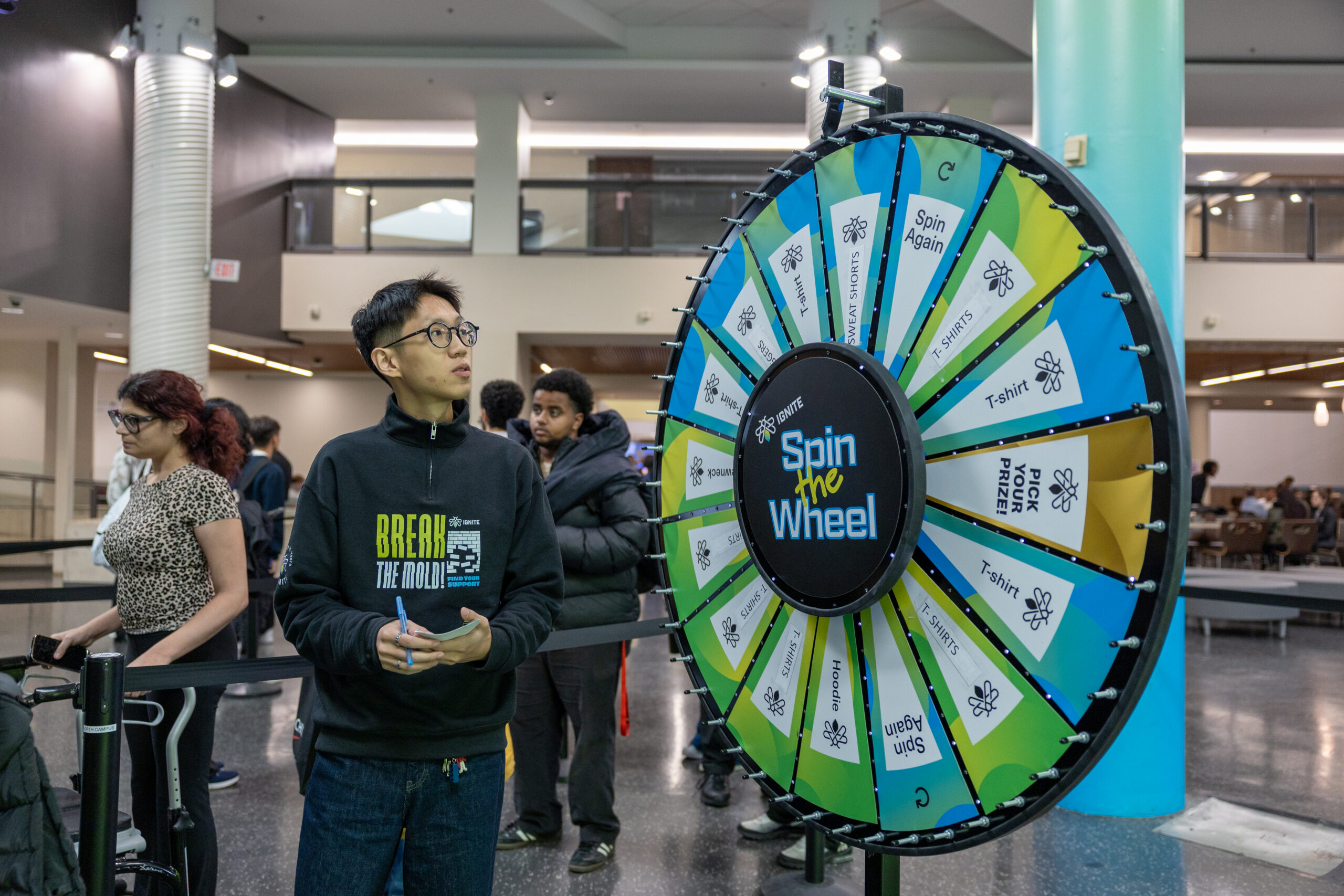 A person stands in front of a colourful "Spin the Wheel" game, holding a notebook and a pen.