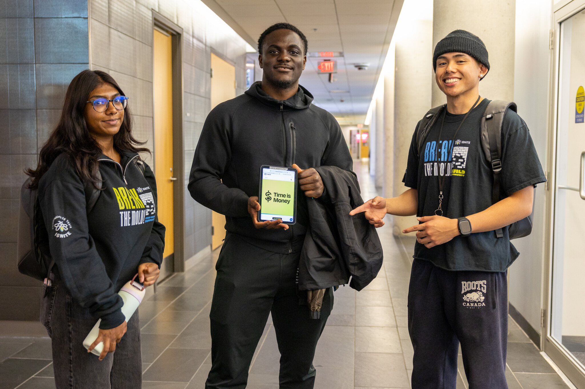 Three students stand in a hallway. The person in the middle holds a phone displaying Time is Money. The other two smile, one pointing at the phone. All wear backpacks and casual clothes.