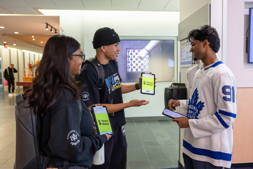 Three students holding tablets with 9 Times or More on the screens talk with another student wearing a white Toronto Maple Leafs jersey in a school hallway.