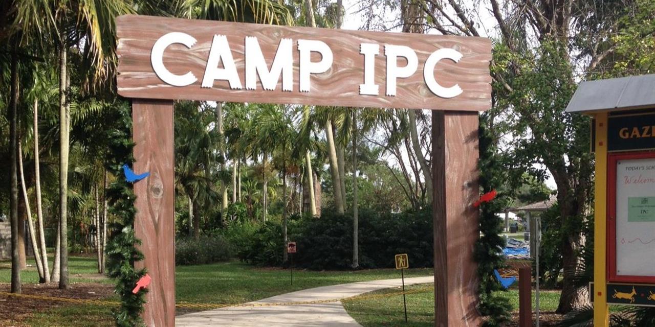 A wooden archway sign reading CAMP IPC stands at the entrance to a park area with palm trees and a sidewalk, decorated with garland and colorful birds.