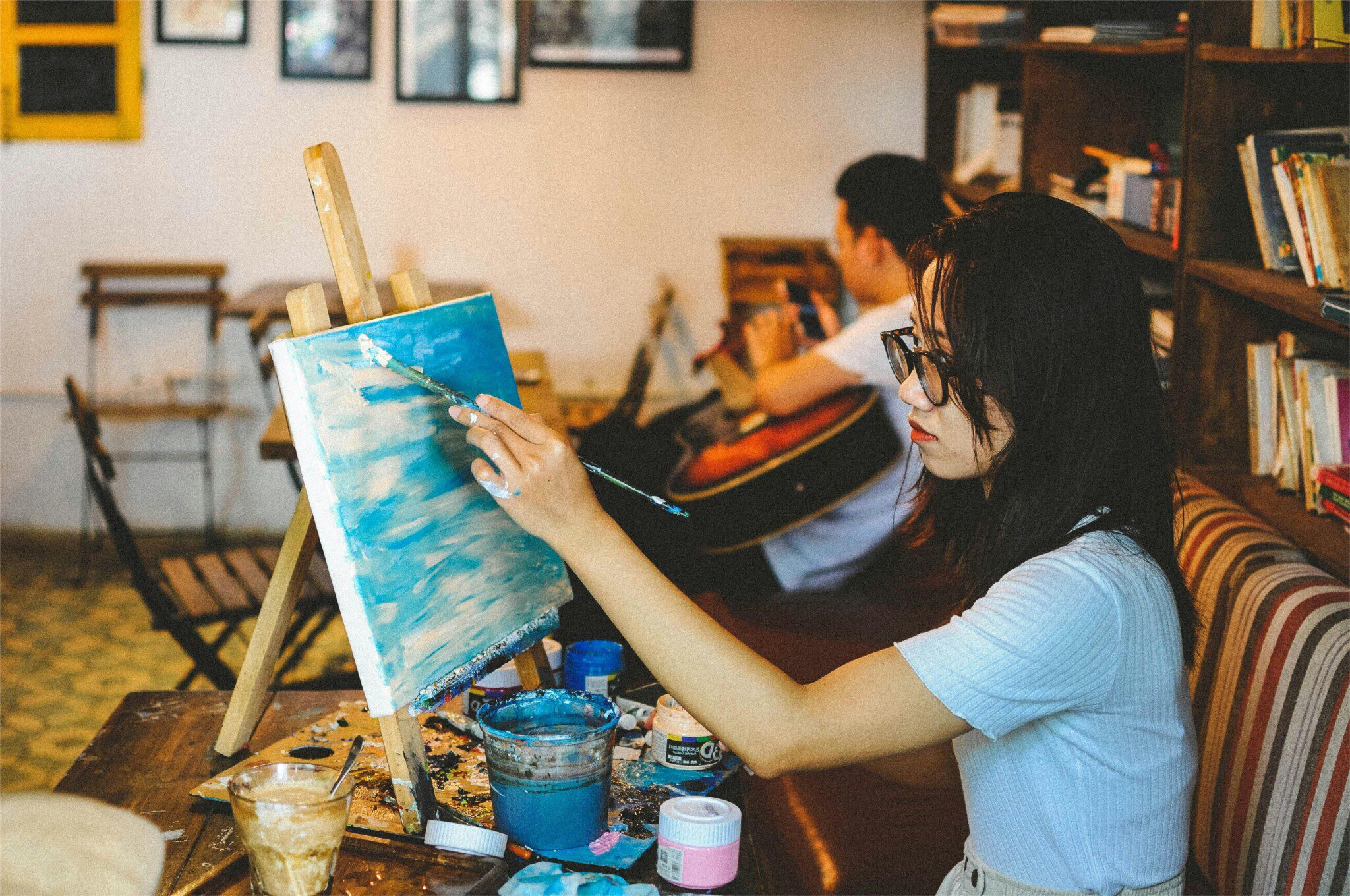 A woman wearing glasses applying blue paint to a canvas on an easel inside a cozy room with bookshelves.