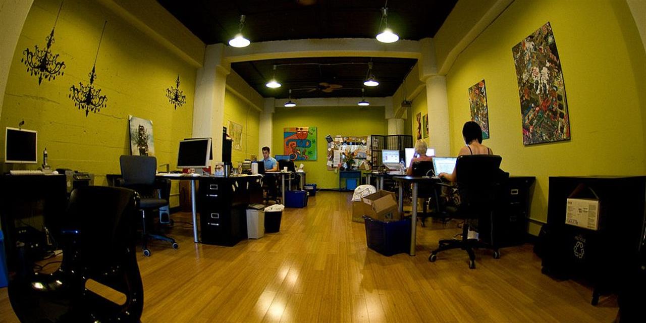 Modern office with bright green walls, wooden floor, several desks with computers, a few people working, eclectic wall art, ceiling lights, and scattered office supplies.
