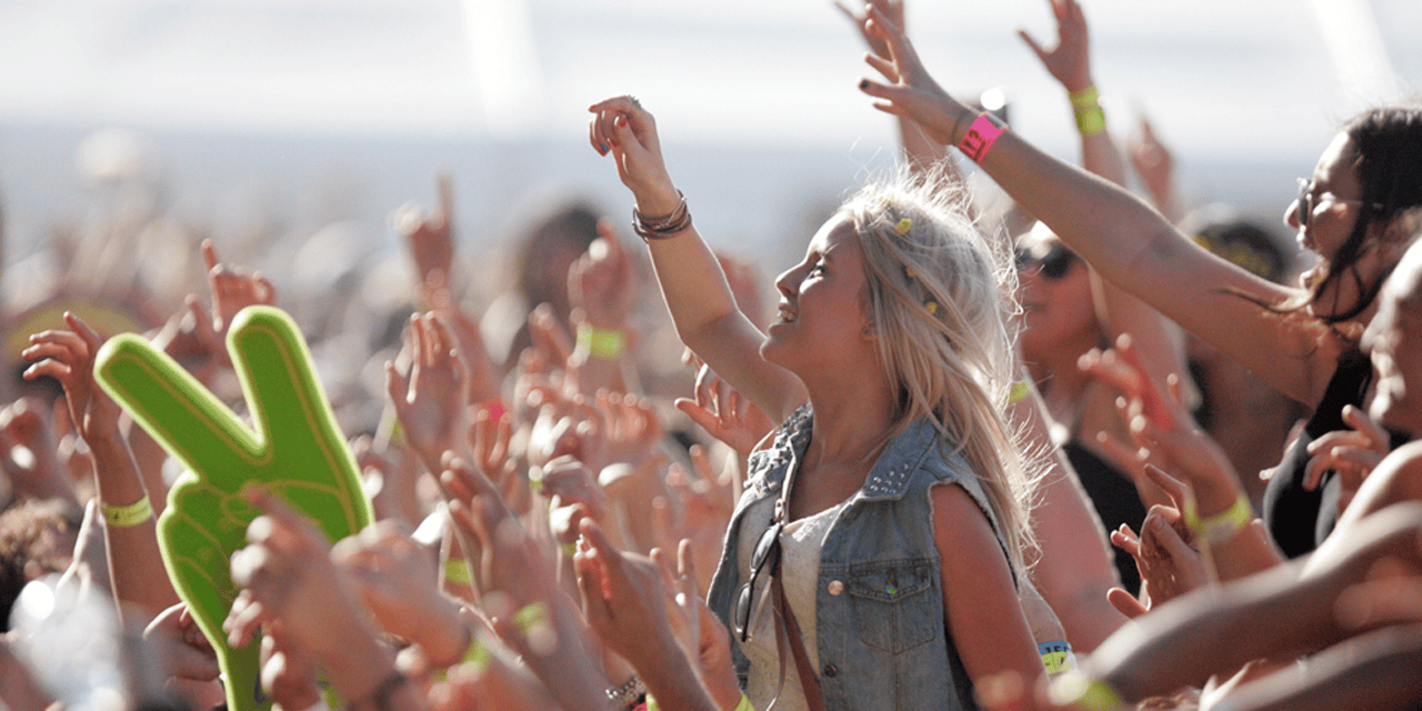 A crowd of people at an outdoor concert, raising their hands and cheering. A woman with long blonde hair in a denim vest stands out in the center, and a green foam hand forms a peace sign on the left. The atmosphere is lively and energetic.
