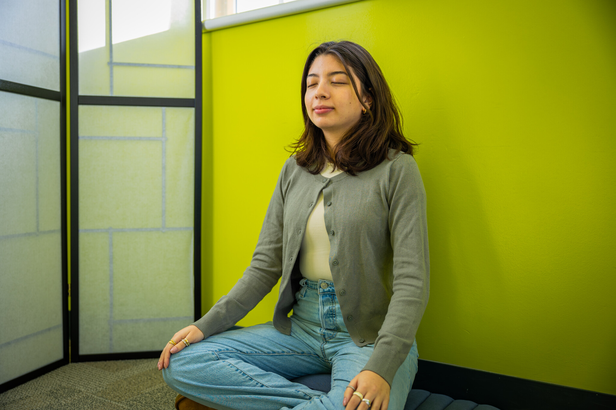 A person with shoulder-length dark hair sits cross-legged on a cushion, eyes closed, and hands resting on knees, meditating against a bright yellow-green wall in a calm indoor space.
