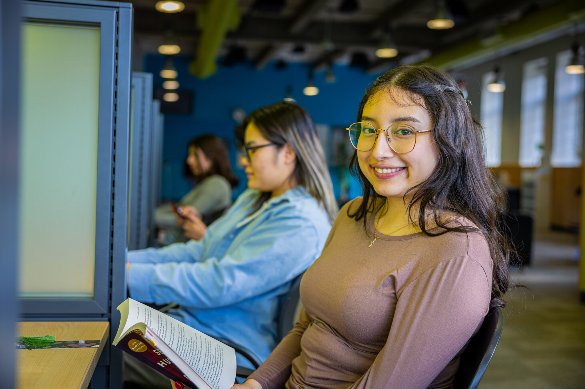 A young woman with glasses smiles at the camera while holding an open book at a study cubicle. Another person sits beside her, focused on their phone. The setting is a modern, well-lit study area.