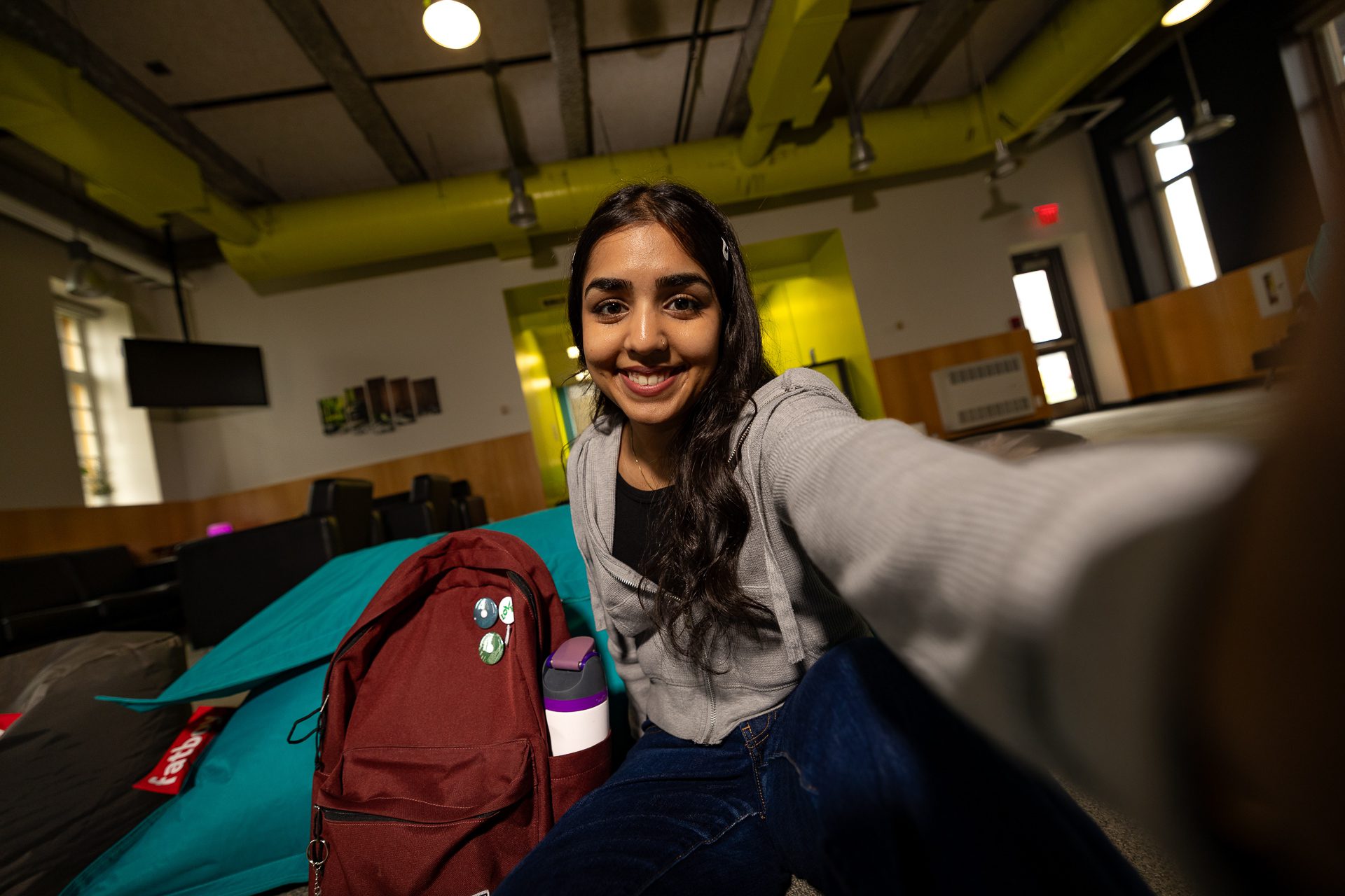 A young woman with long dark hair smiles at the camera, sitting indoors with a maroon backpack and a water bottle beside her, in a brightly lit room with green and yellow accents.