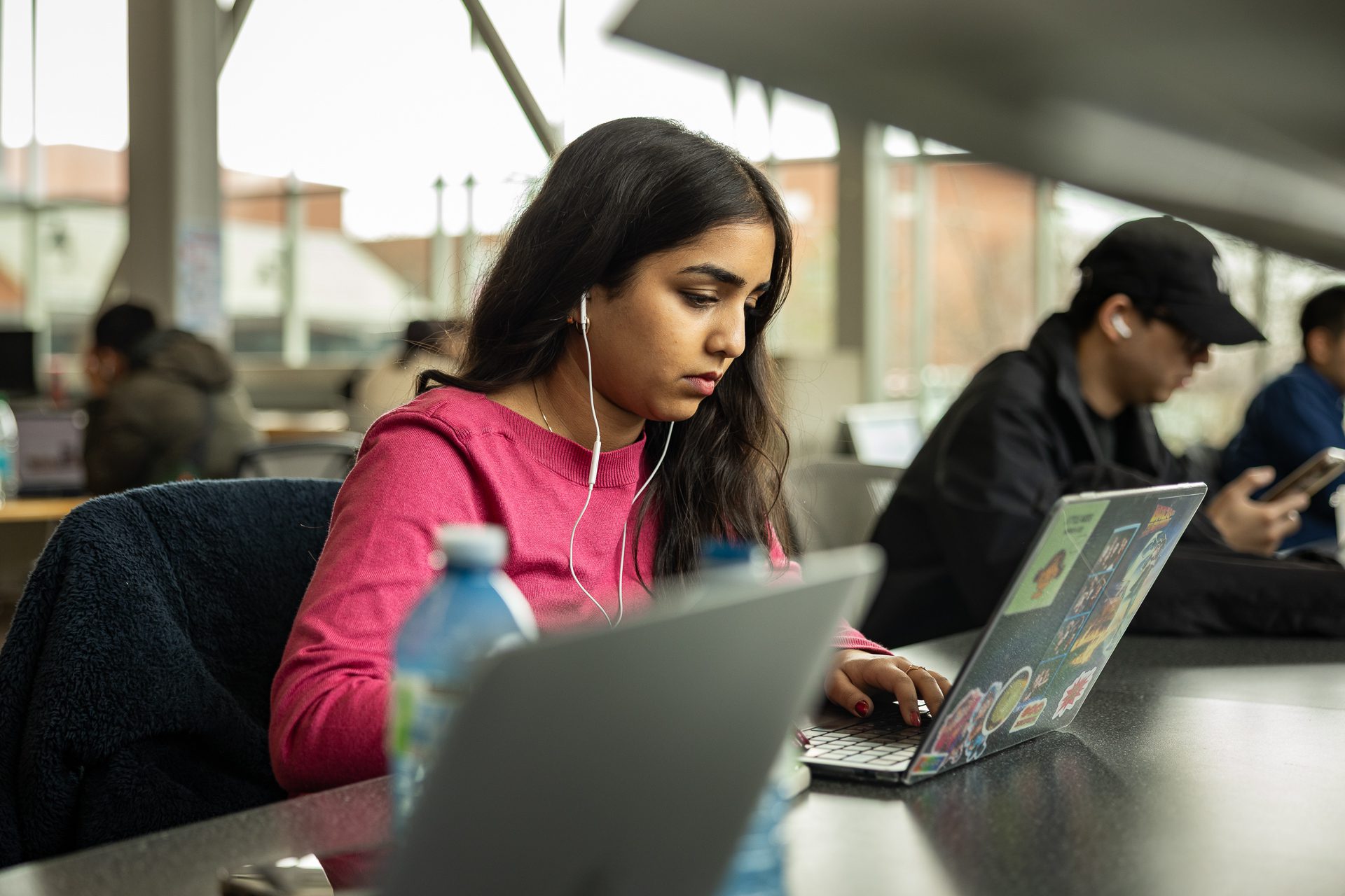 A student wearing earphones typing on a sticker-decorated laptop at a shared study table.