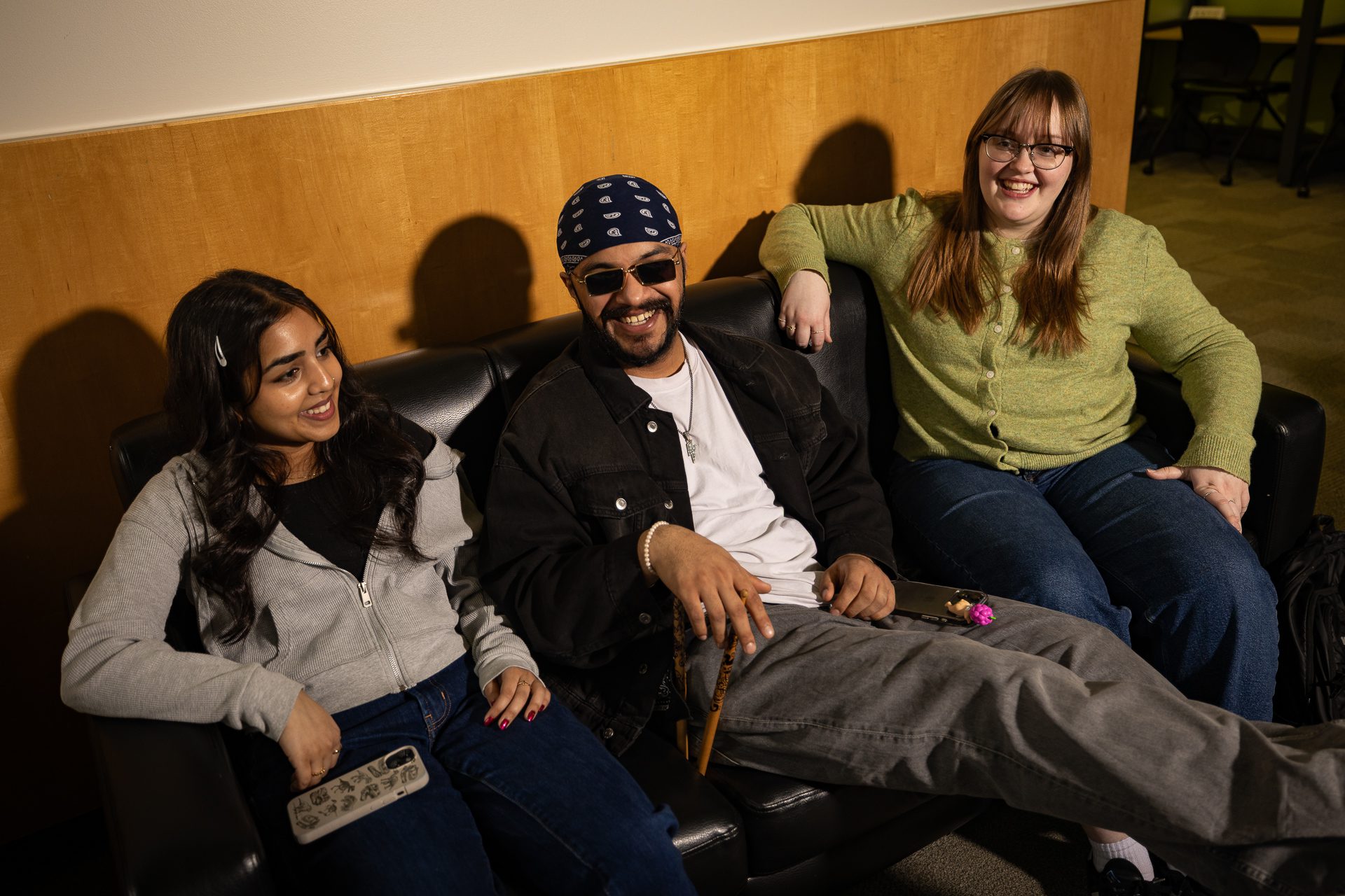 Three people sit closely together on a black couch, smiling and laughing. The person in the middle wears sunglasses and a bandana, while the two people beside them wear casual clothes and glasses. They appear relaxed and happy.
