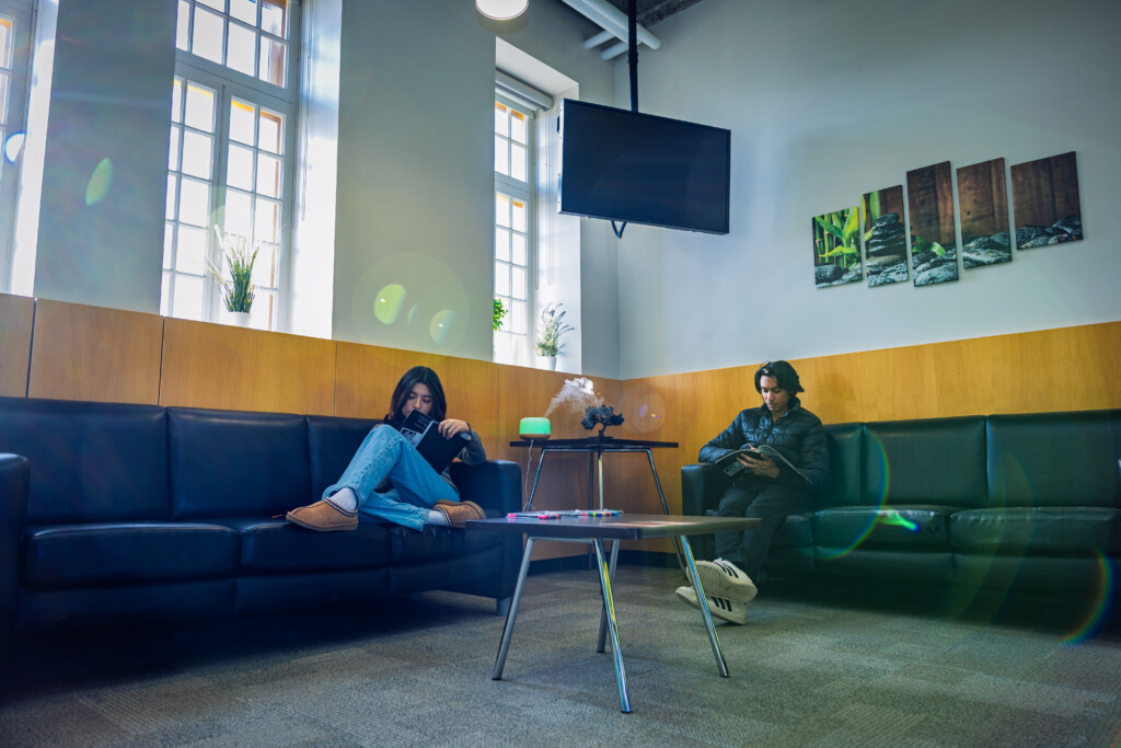 Two students reading books while sitting on leather couches in a quiet, sunlit lounge area.