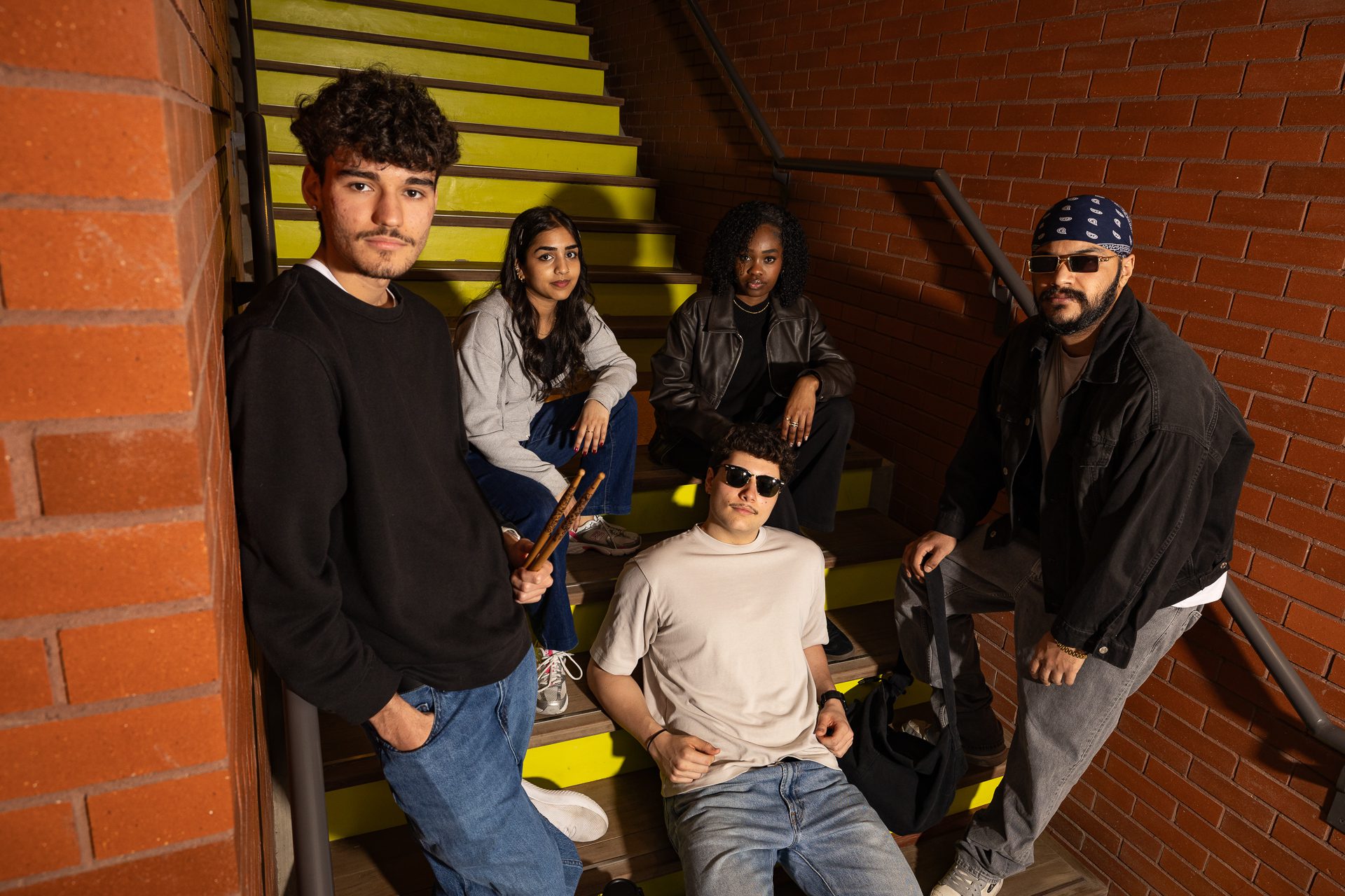 Five young adults sit and stand on yellow stairs next to a brick wall, posing for the camera. They have diverse appearances and casual clothing, with one person wearing sunglasses and another with a bandana.
