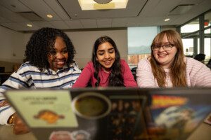 Three smiling students gathered around a table looking together at a laptop screen.