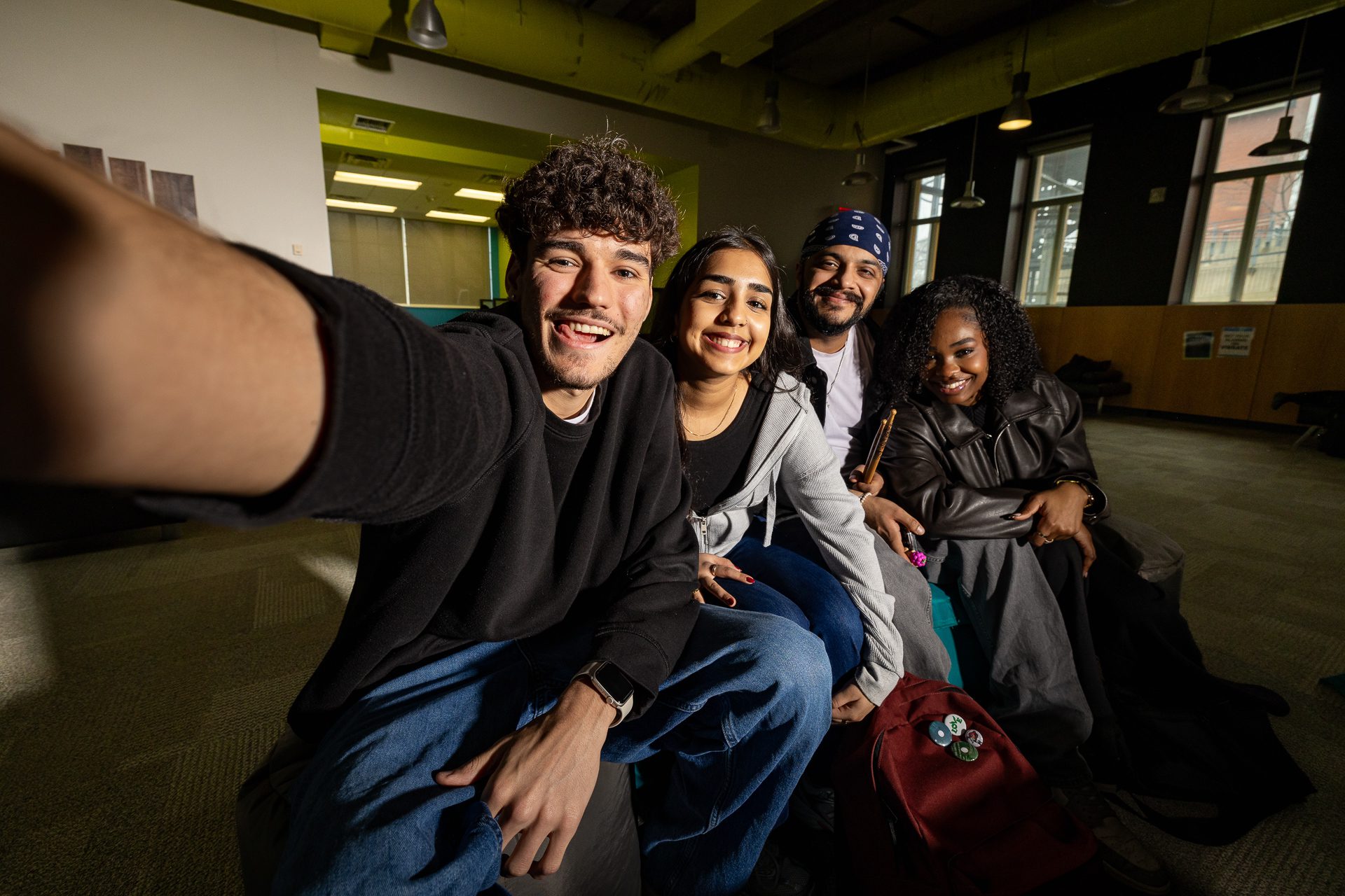 Four young adults sit close together indoors, smiling at the camera as one of them takes a selfie. The group appears happy and relaxed in a modern, well-lit room.