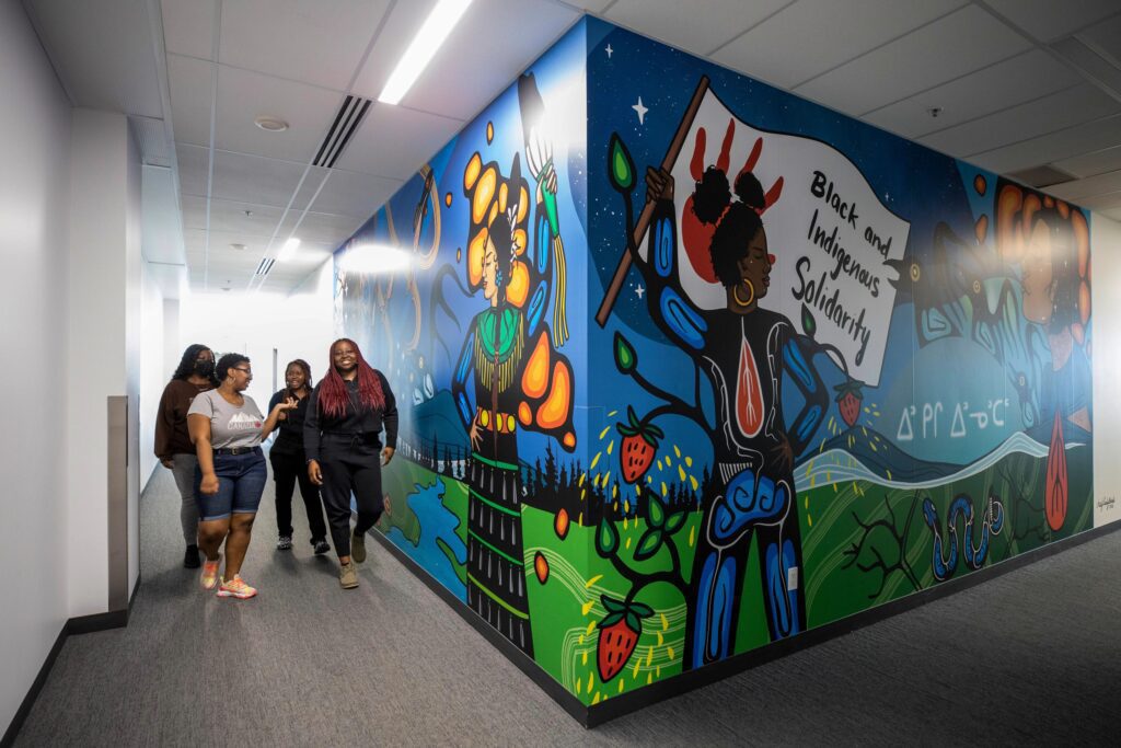 A group of women are walking beside a wall covered in Indigenous artwork.