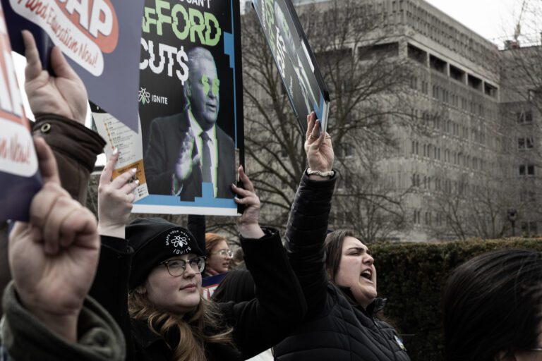 Crowd of protesters outdoors holding up political signs, including a blue poster reading 'FORD CUTS,' as people chant in a city setting.