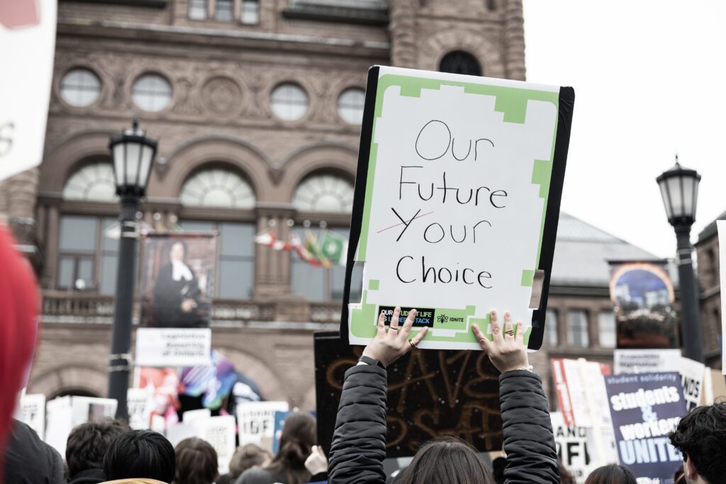 A person holds a sign that reads "Our Future Your Choice" at a protest in front of Queen’s Park with other demonstrators visible.