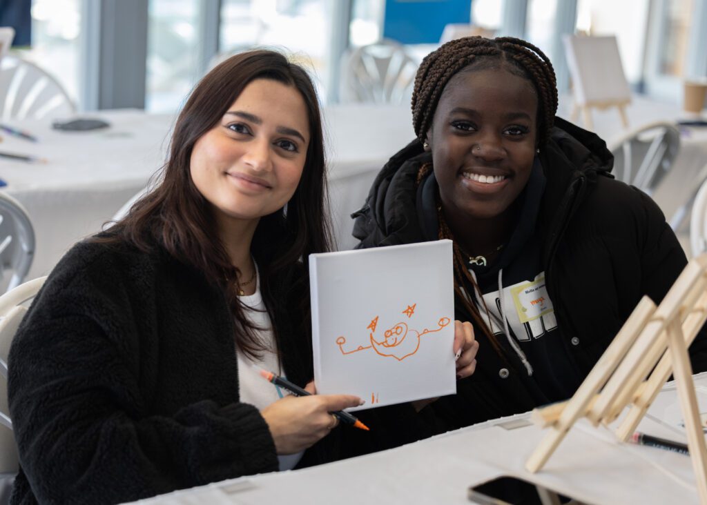 Two women sit at a table. One holds up a small whiteboard with an orange cartoon drawing on it.