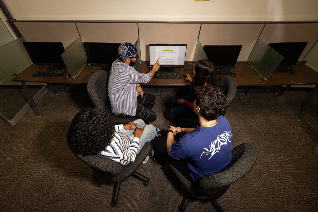 Four students in a circle all looking at a desktop computer in a workspace. One student points at the text on the computer while another holds the mouse. 